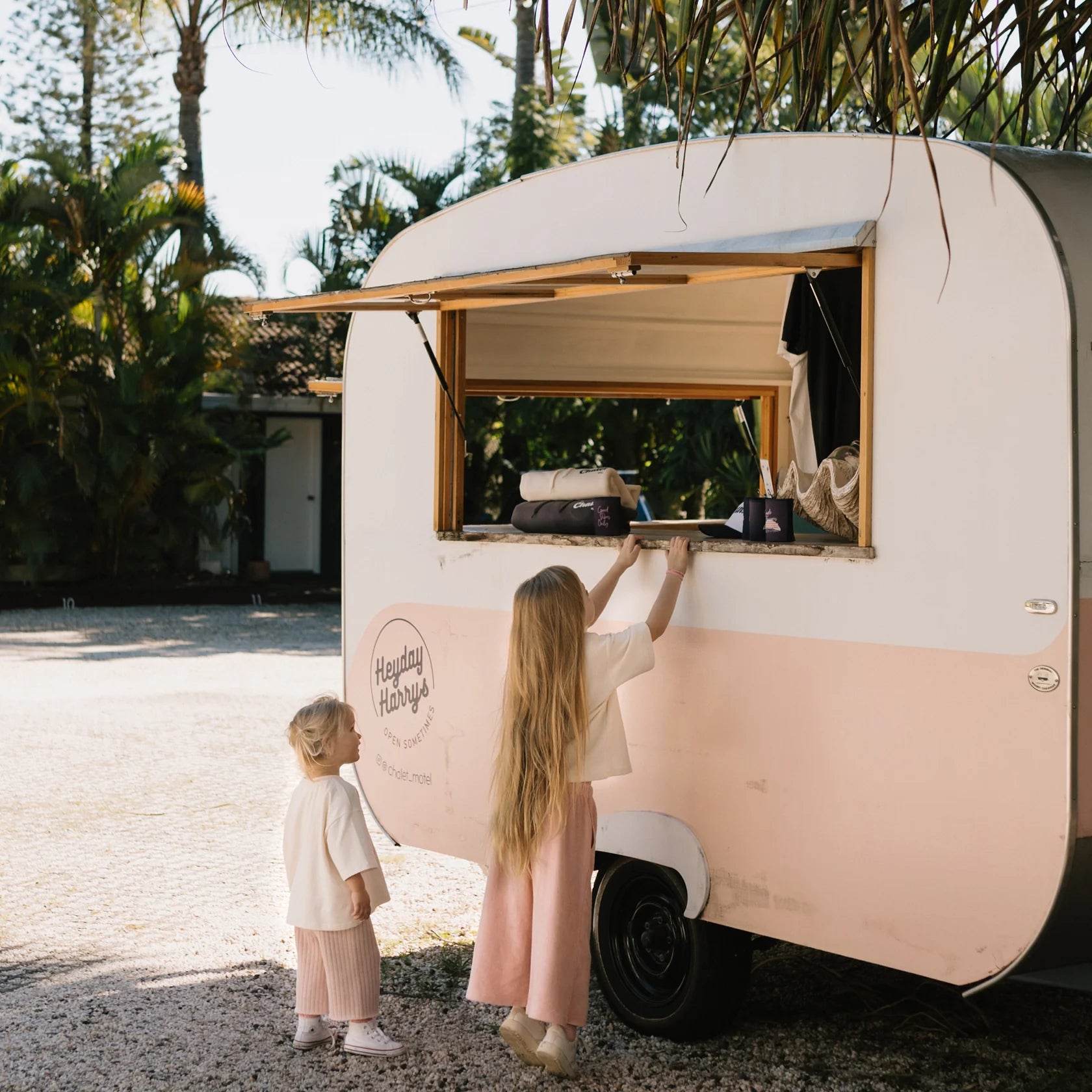 Two children with long hair stand beside a white and pink food truck named Happy Harry's. One child, wearing GOLDEN CHILDREN's Confetti Wide Leg Pants in Pink Parfait, reaches up to the serving window, while the other child, dressed similarly in soft terry towel cotton, looks on. The background features palm trees and a clear sky.