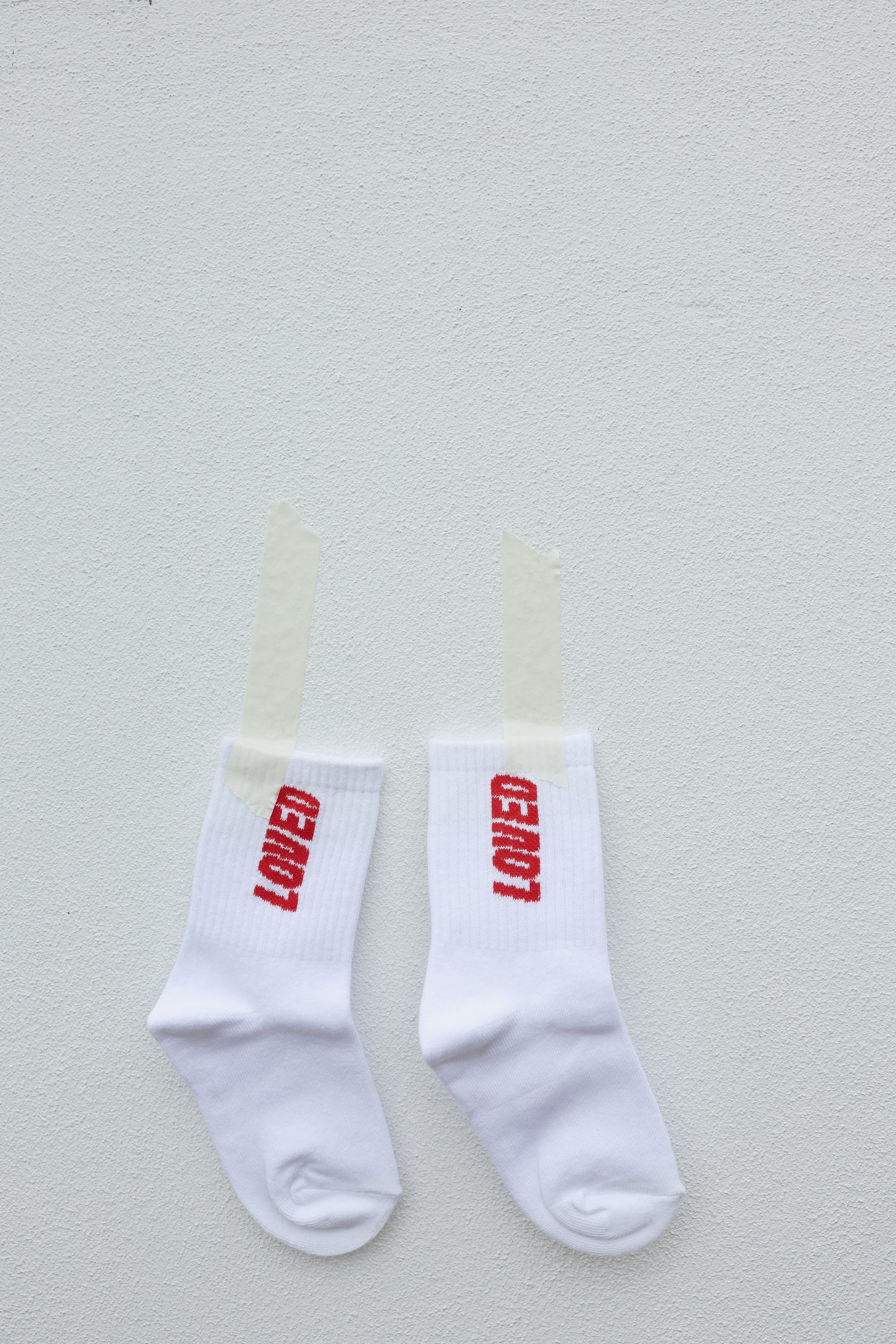 A pair of TINY LOVE CLUB's Loved Socks is taped to a white wall with transparent tape. Each sock has the word "LOVED" written in red, vertically across the ankle, and features elastic banded ankles for a snug fit.