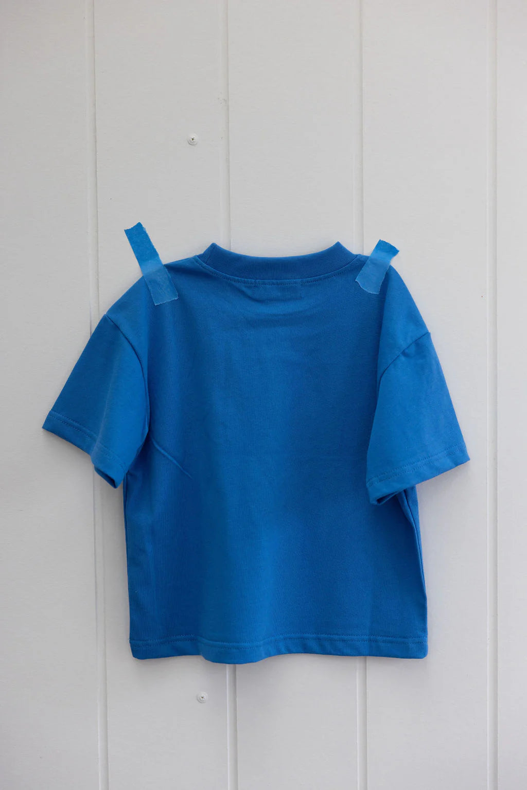 The Sunday Siblings Il Sole Tee Blue/Yellow, made from soft cotton fabric, is taped with blue tape to a vertically paneled white wall. The simple yet elegant design of this t-shirt stands out against the grooved backdrop.