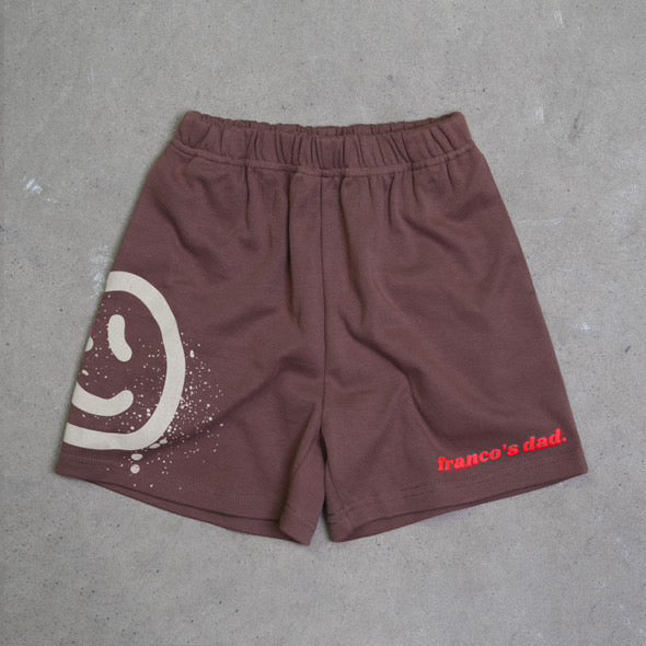 Acid Dye Shorts Brown by FRANCO'S DAD on a gray surface, featuring a large white smiley face and red "franco's dad" text. Made from stretch cotton, these shorts have an elastic waistband for casual comfort, ideal for lounging in style.