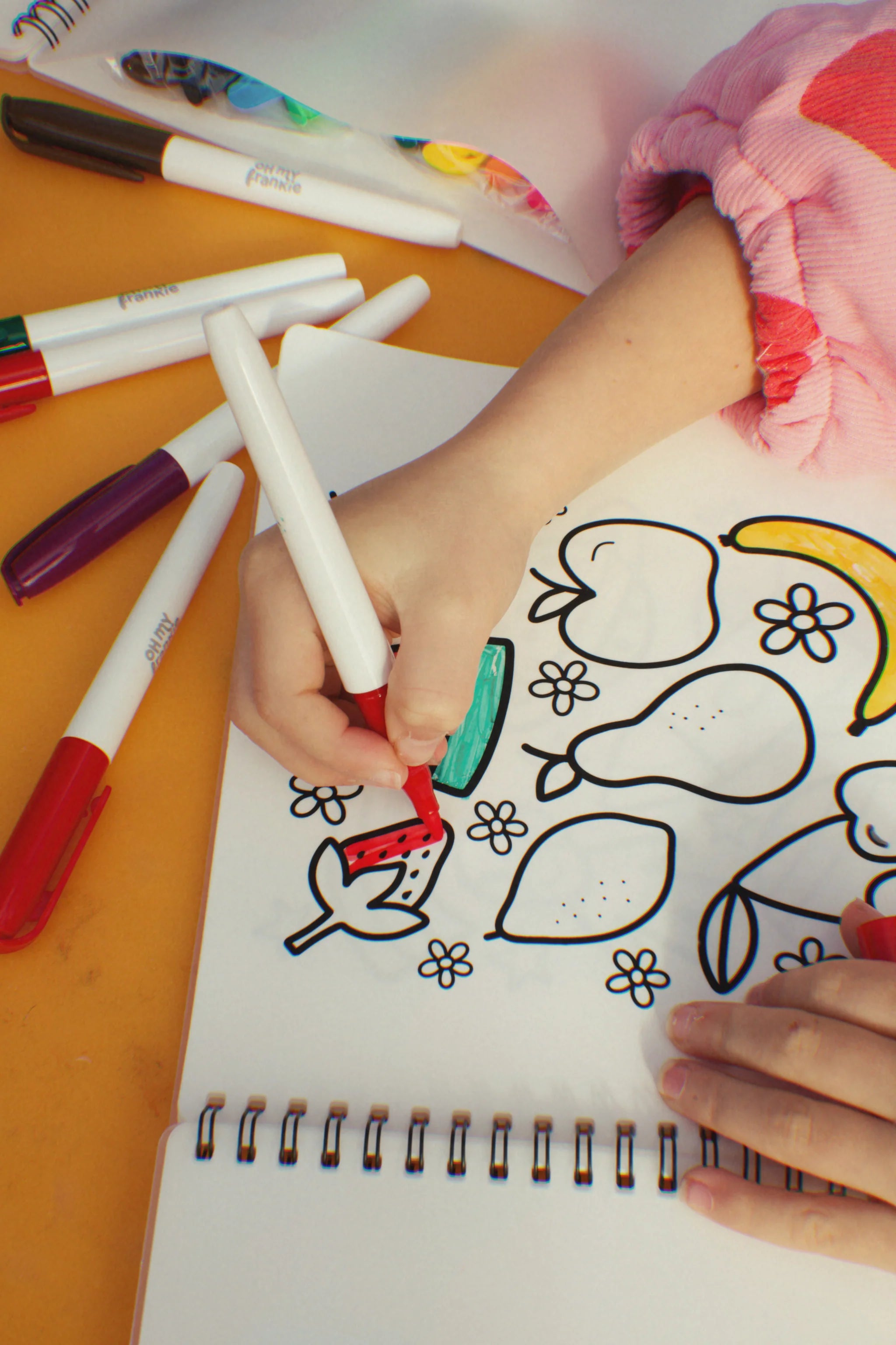 A child in a pink long-sleeve shirt creatively colors a fruit-themed page in the Oh My Frankie ~ Oh My Scribbles Reusable Colouring Book. Using a red non-toxic marker, surrounded by other colored markers on a yellow surface, the activity highlights kids' artistic expression.