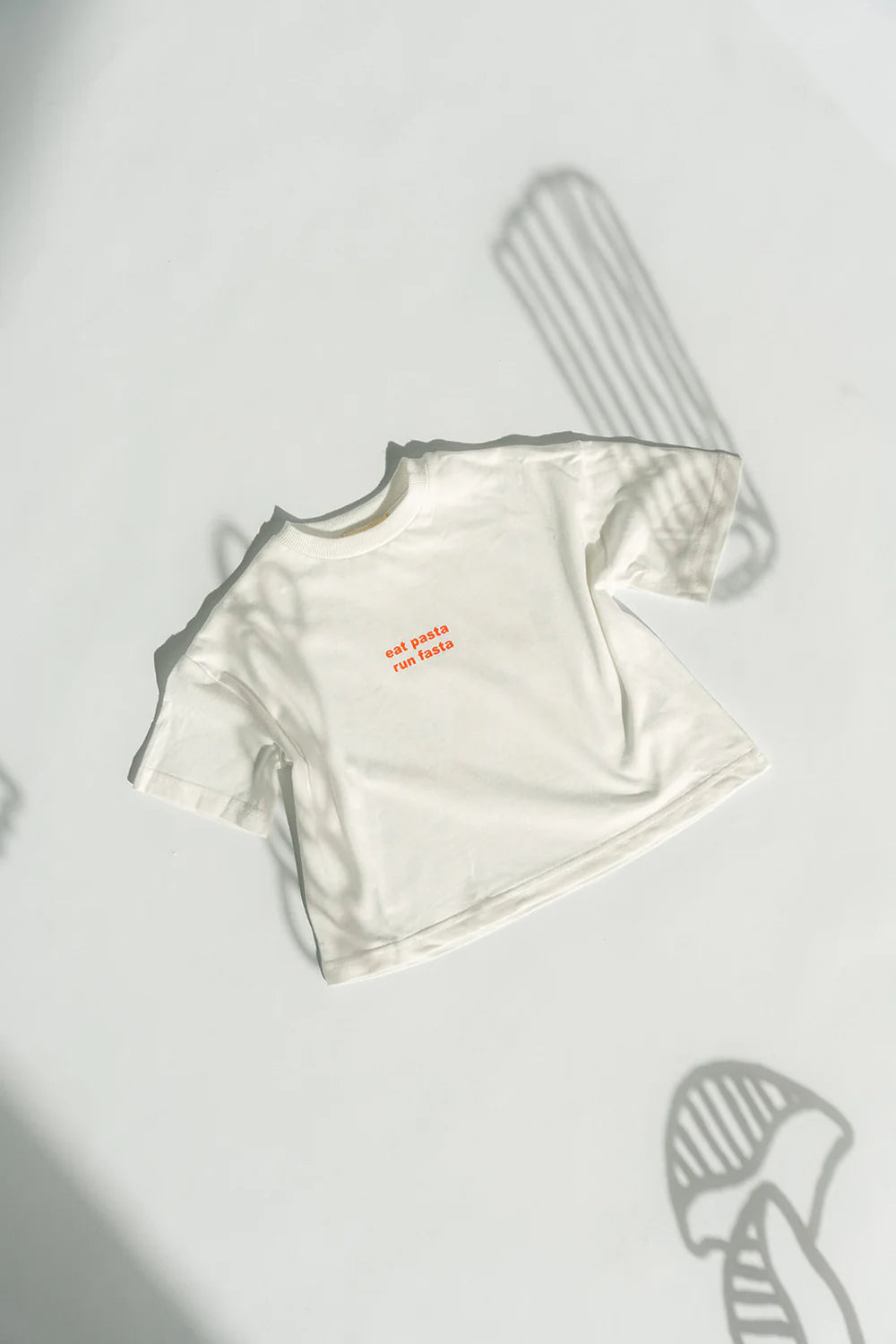 The Pasta Tee White from LENN LABEL, a kids' short-sleeved cotton jersey shirt, lies flat on a light background. The garment features small red text in the center of the chest area. Shadows of abstract shapes and objects are cast around it.