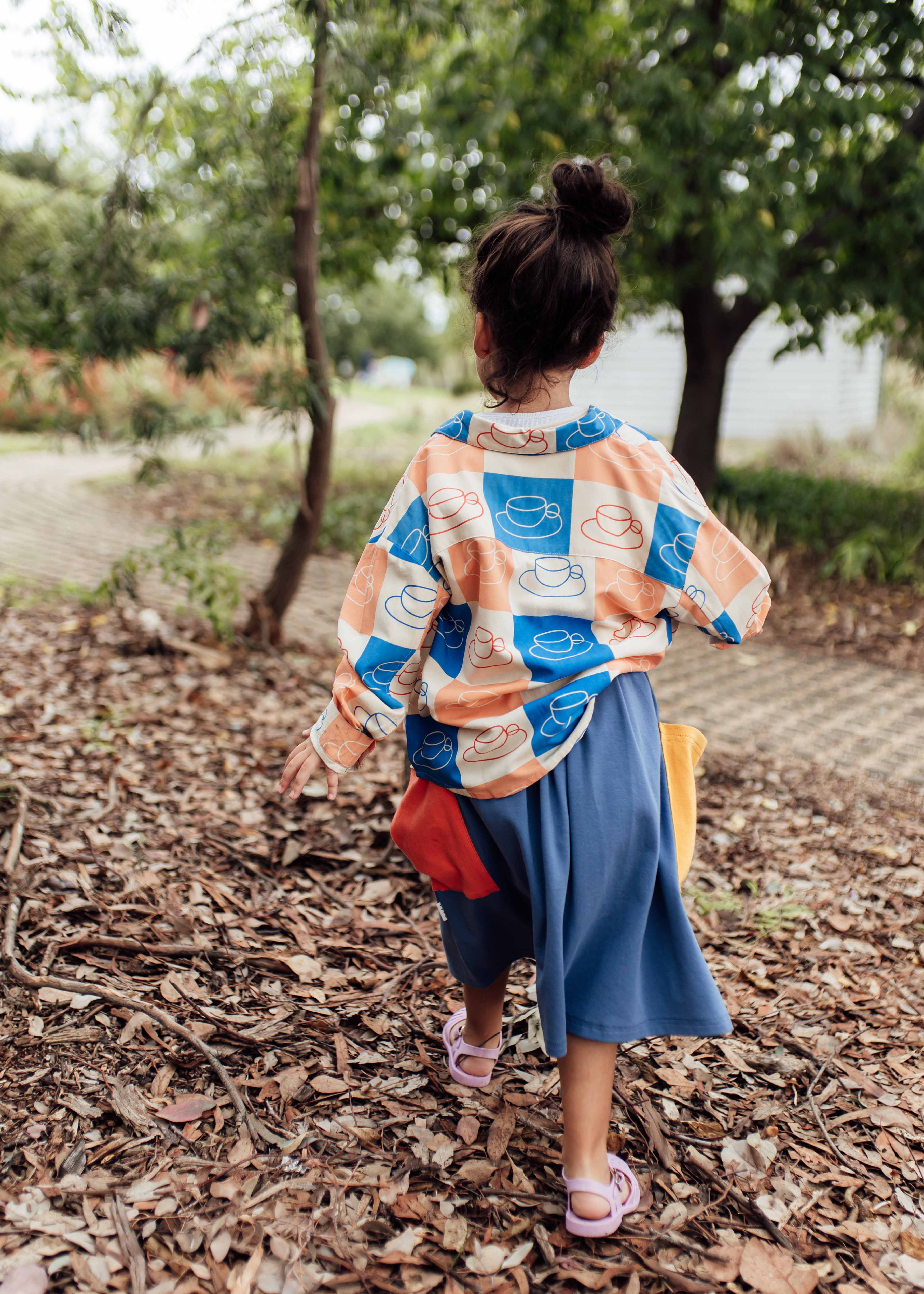 A child with a bun hairstyle walks away on a leaf-covered path in a wooded area. They are wearing the Cups Button Down Shirt, designed by OLIVE + THE CAPTAIN, made from a premium cotton linen blend, paired with blue pants. Trees and greenery are visible in the background, adding to the picturesque Melbourne-designed scene.