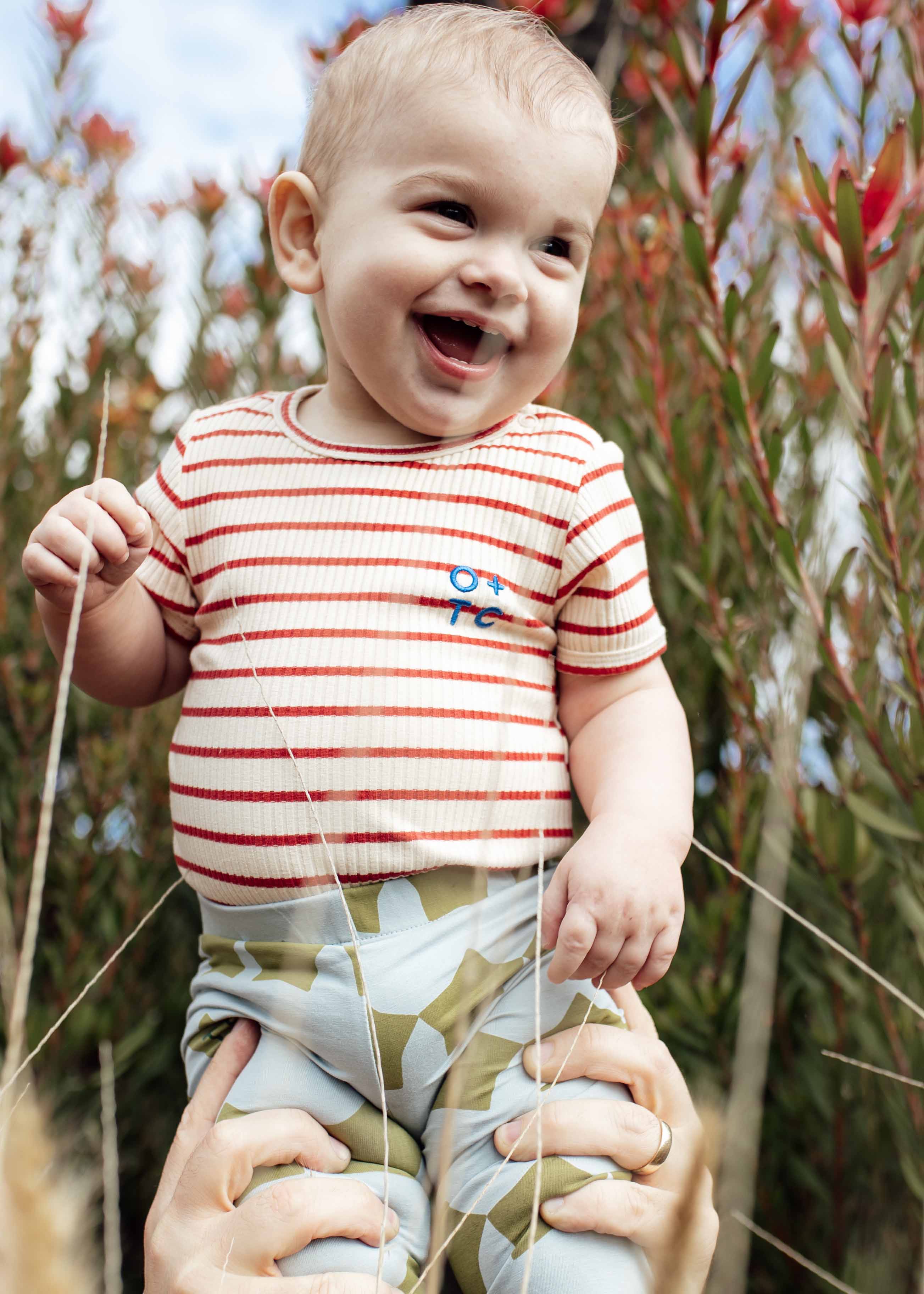 A smiling baby wearing the Samba Stripe Bodysuit from OLIVE + THE CAPTAIN, made of premium cotton and featuring red and white stripes, along with blue-green patterned pants is being held up by two adult hands. The background showcases tall green foliage with red-tipped leaves against a clear sky.