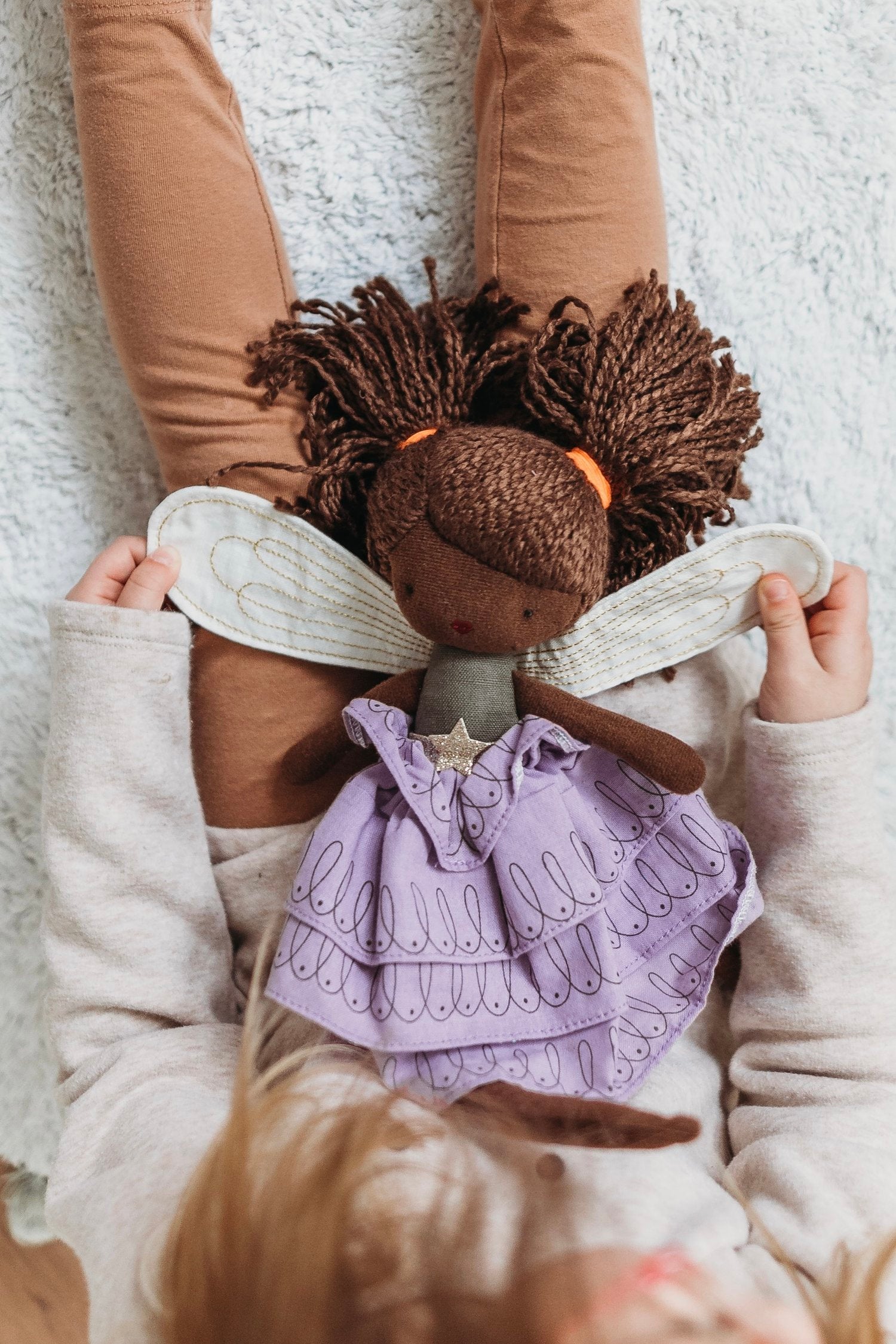 A child sitting on the floor holds a dark-skinned PICCA LOULOU Fairy Feline 35cm rag doll with curly hair tied in pigtails and adorned with orange hair ties. The Fairy Feline doll wears a purple dress with white wing-like attachments on its back. The child wears light-colored clothes.