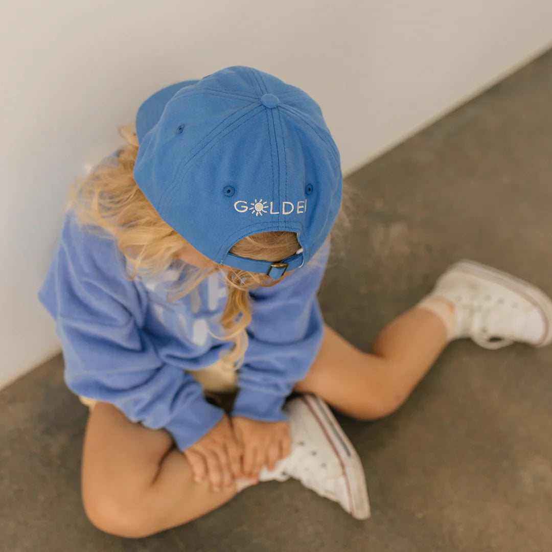 A child sits on a concrete floor, wearing a Lobster Cap Deep Sea Blue from GOLDEN CHILDREN embroidered with "GOLDEN" on it in reverse. This trendy dad cap includes an adjustable brass clasp. The child is dressed in a light blue sweatshirt, beige shorts, and white sneakers, with their hands clasped together.