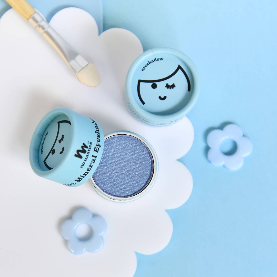 A container of no nasties kids' **Kids Eco Natural Pressed Eyeshadow & Blush** in blue sits open on a cloud-shaped white surface, featuring a decorative face design on the lid. Nearby, a small applicator brush and flower-shaped ornaments are arranged against a blue background. This cruelty-free product is made with natural ingredients for gentle application.