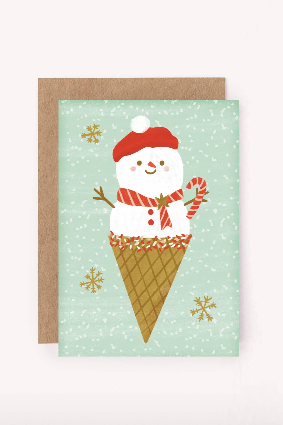 The Ms Snow Cone Mini Card from LAUREN SISSONS STUDIO showcases an adorable design of a snowman sporting a red hat and scarf, delightfully perched in a waffle cone while holding a candy cane among falling snowflakes on a light green background. Crafted from recycled paper, this charming card comes paired with a brown envelope.