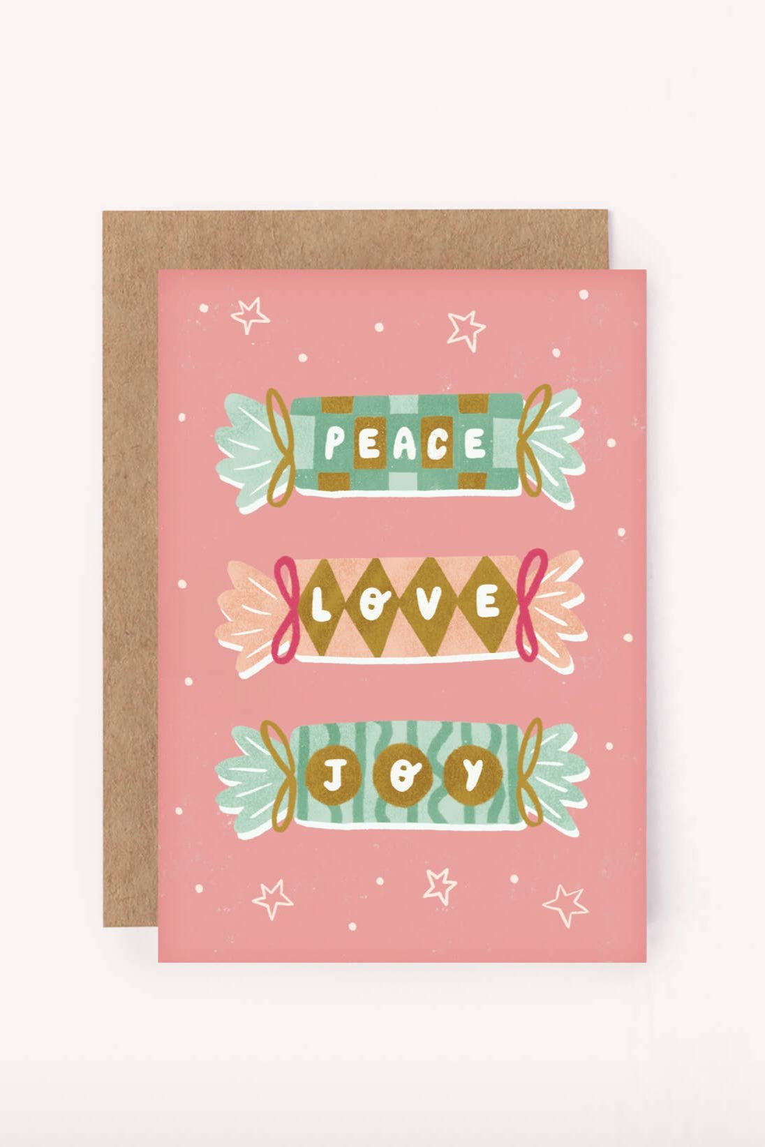 The Peace Love Joy Christmas Crackers Mini Card by LAUREN SISSONS STUDIO features a festive design on pink recycled paper with colorful banners declaring "Peace," "Love," and "Joy," accented with stars and snowflakes, and produced sustainably. It is accompanied by a brown envelope.