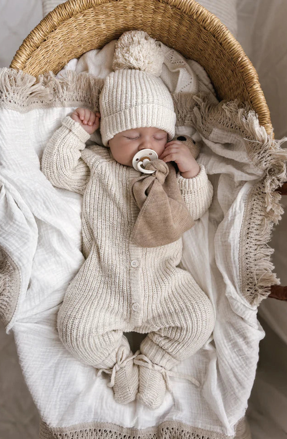 A sleeping baby is nestled in a wicker basket lined with a white, fringed blanket. The baby is dressed in the Classic Knit Romper Honey by ZIGGY LOU and matching booties, complete with a hat. With a pacifier in their mouth, the scene is cozy and serene, capturing the essence of tranquil winter outfit charm.