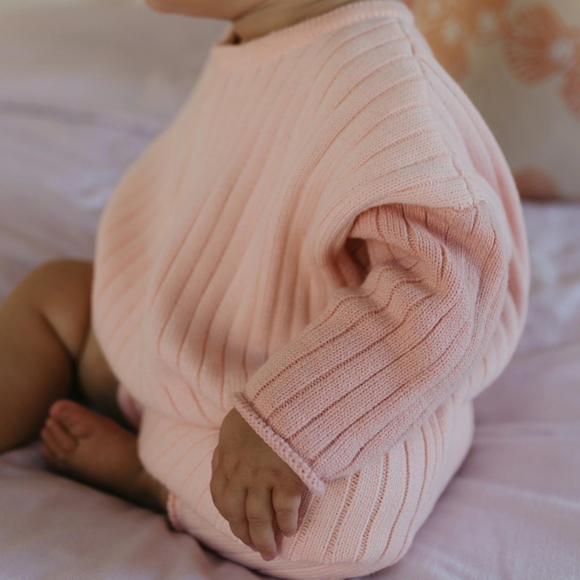 An infant dressed in a cozy, ribbed pink Golden Knit Romper Strawberry Sundae by GOLDEN CHILDREN is seated on a soft surface, with only part of the face and body visible. The romper’s texture is clearly defined, made from 100% cotton knit, and the background suggests a calm, indoor setting.