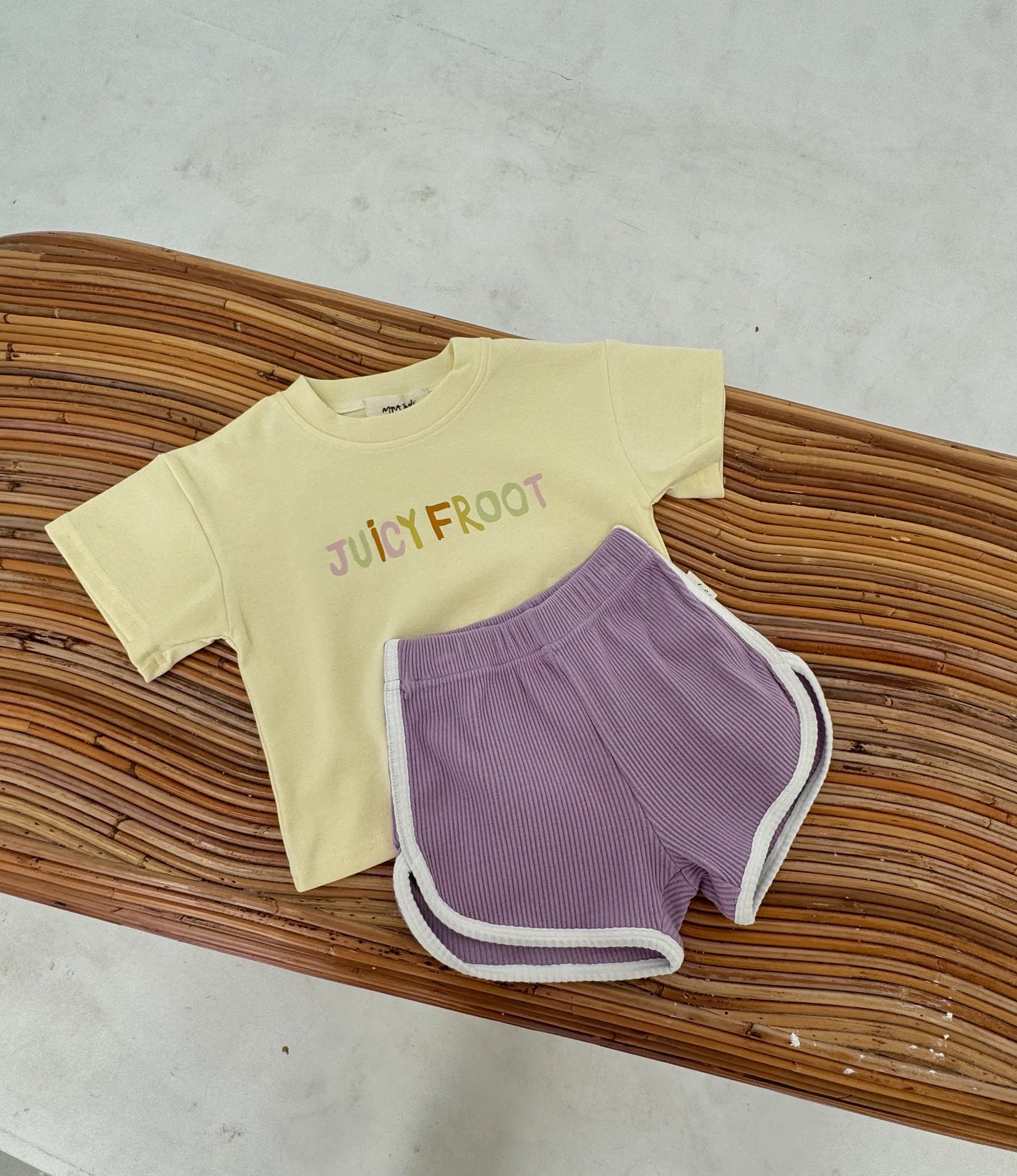 A cozy oversized Juicy Froot Tee in yellow, featuring the bold "JUICY FROOT" design, is perfectly complemented by MYLA JANE's Contrast Rib Shorts Grape in a vibrant purple with white trim. This chic outfit is artfully arranged on a wooden bench against a softly lit background.