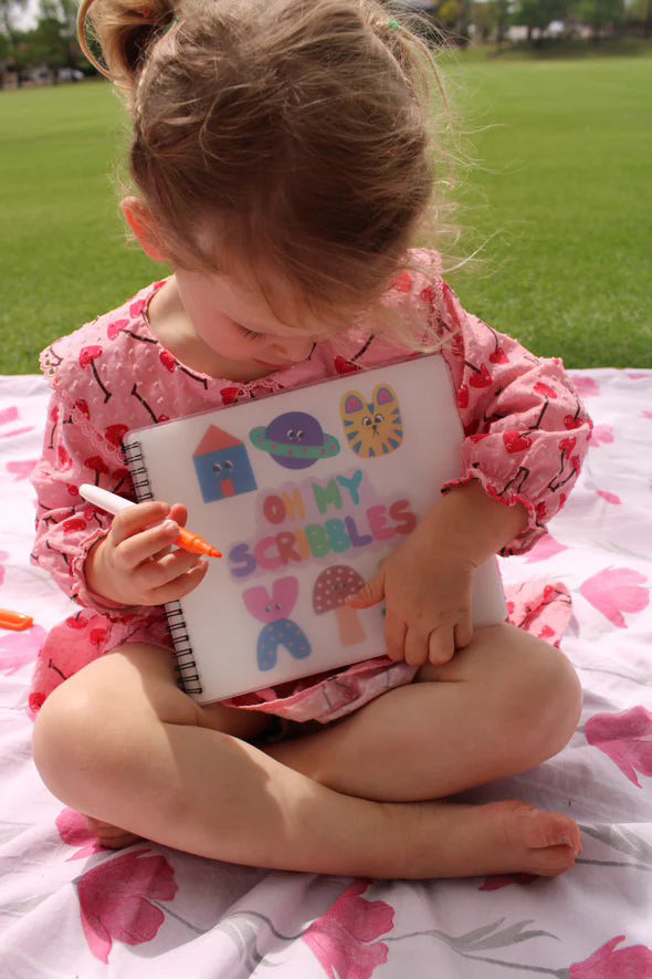 A young child in a pink dress sits on a blanket in the grass, holding an "Oh My Frankie ~ Oh My Scribbles" reusable coloring book. She draws with non-toxic markers, her focus reflecting pure kids' creativity.