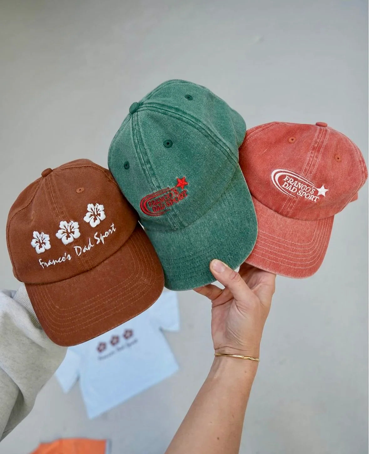 A person holds up three PRE-ORDER Sports Caps by FRANCO'S DAD in brown, green, and rust. The brown cap is detailed with white embroidered flowers and text. The green cap, enhanced with a red acid dye, displays a striking red star and text. The rust cap also features a star design. A blurred t-shirt is visible in the background.