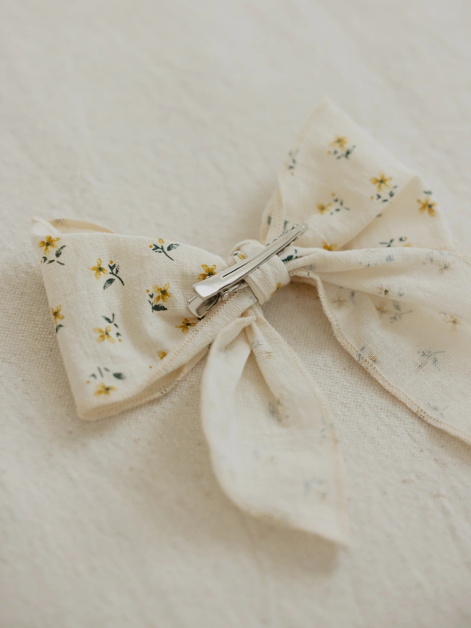 A close-up of the VALENCIA BYRON BAY Cotton Bow Marigold, a charming handmade hair accessory featuring a vibrant floral pattern with small yellow marigolds and green leaves. This delightful bow is secured with a metallic clip on a light beige textured surface, making it an ideal gift.