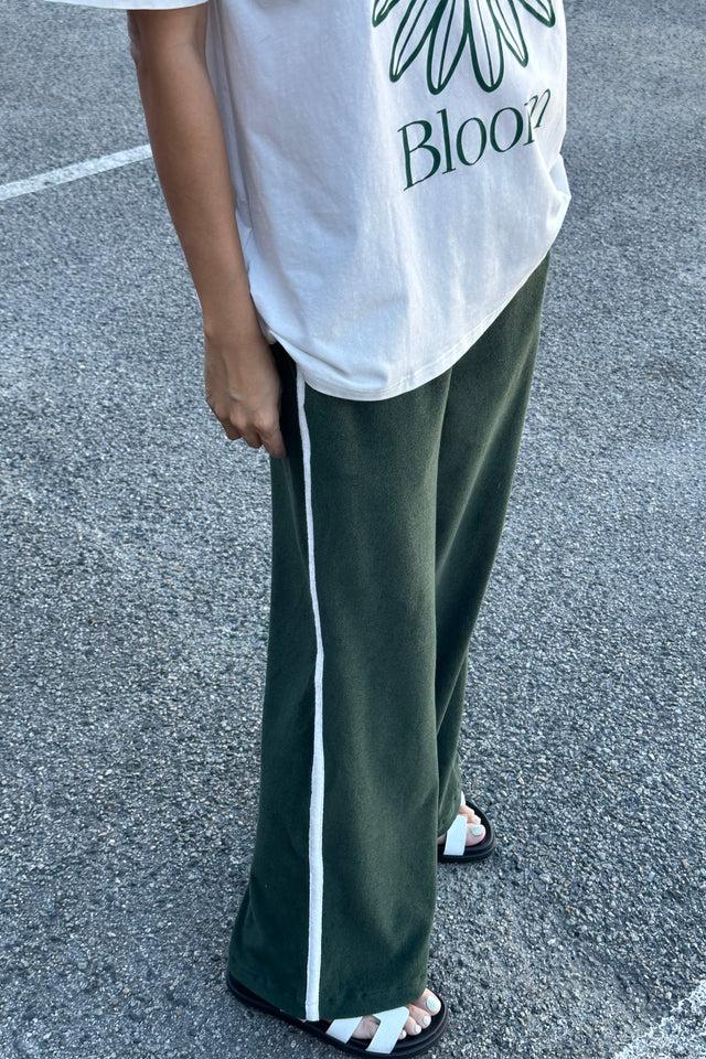 A person is standing on a paved surface wearing a white T-shirt with a green floral design and the partially visible word "Bloom," paired with TINY TROVE's Piper Racer Pants Moss featuring white vertical stripes and an elasticated waistband, along with white sandals. Only the person's upper torso and legs are visible in the image.