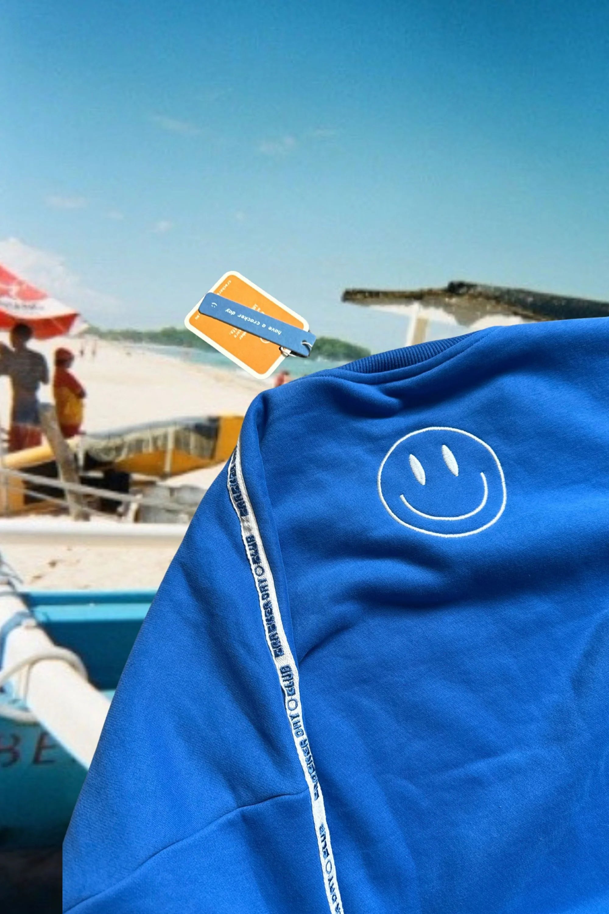 The Cracker Day Club - Cloud Racer Sweater Blue by CRACKER DAY CLUB, featuring a white smiley face and logo stripes, is displayed on a surface with a blue and orange luggage tag. In the background, people are near boats on a sunny beach.