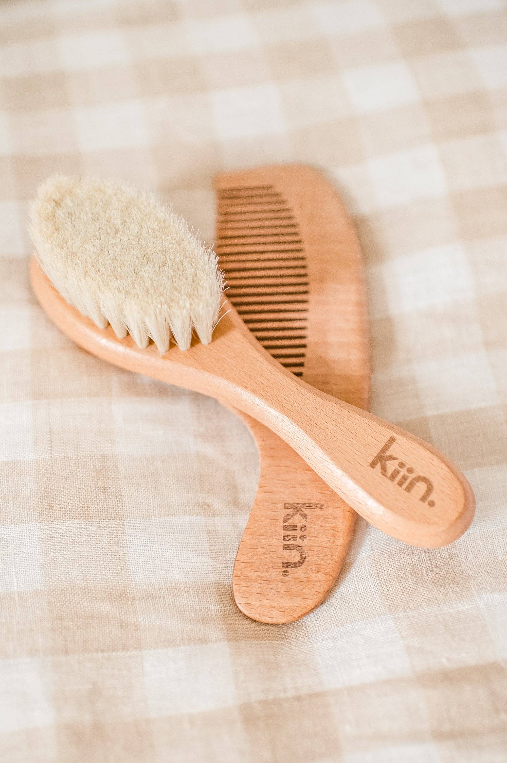 Resting on beige and white checkered fabric, the Wooden Baby Brush and Comb Set by KIIN BABY features a wooden baby hair brush with soft goat wool bristles and an eco-friendly wooden comb, both elegantly marked with "Kiin," embodying natural beauty and gentle care.