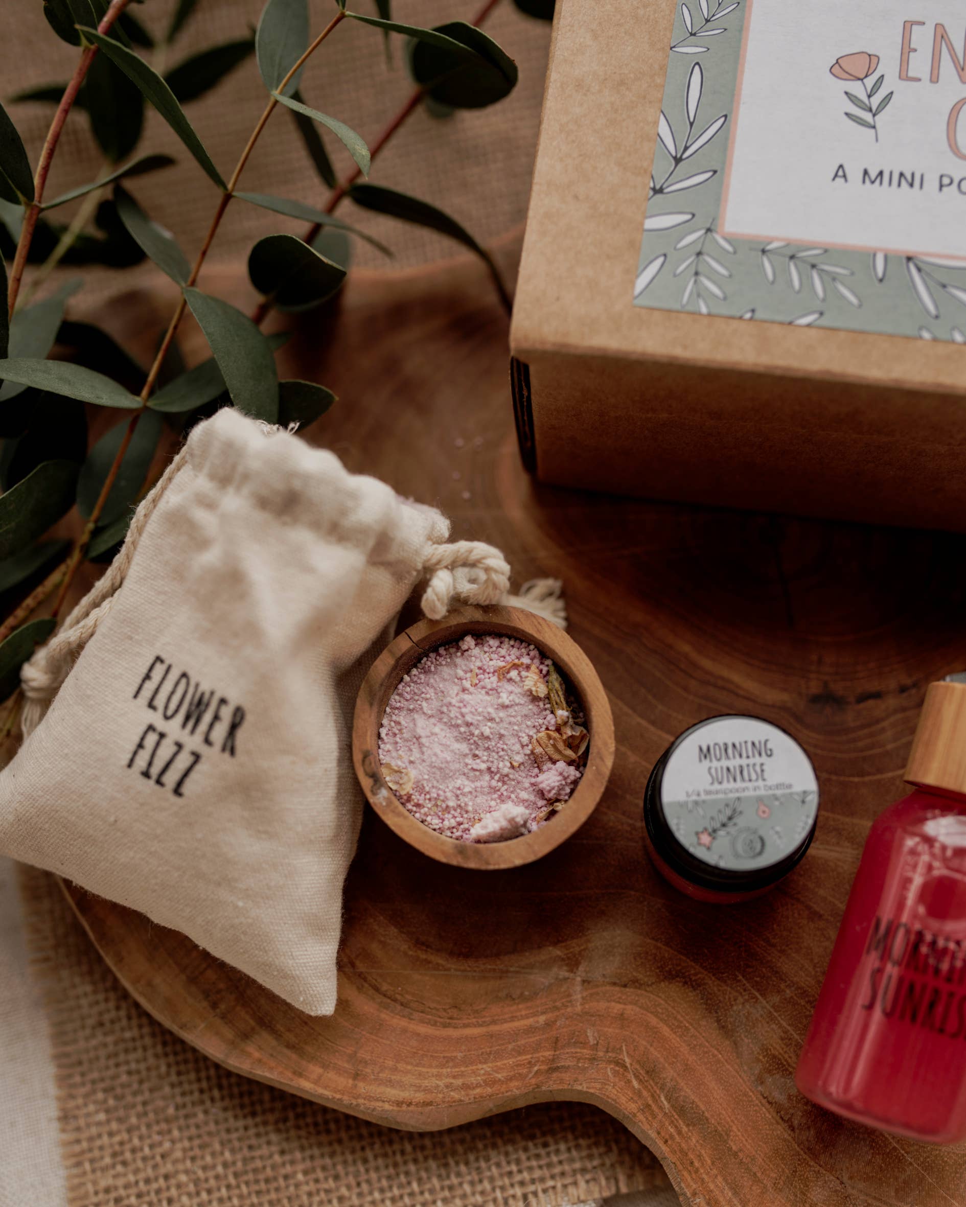 A wooden tray showcases THE LITTLE POTION CO's MINI Enchanted Garden Potion Kit, perfect for sensory play. The kit includes a labeled "Flower Fizz" fabric pouch, a wooden bowl with pink bath salts, a jar marked "Morning Sunrise," and a partially visible red bottled product, all nestled among green leaves and accompanied by a cardboard box.