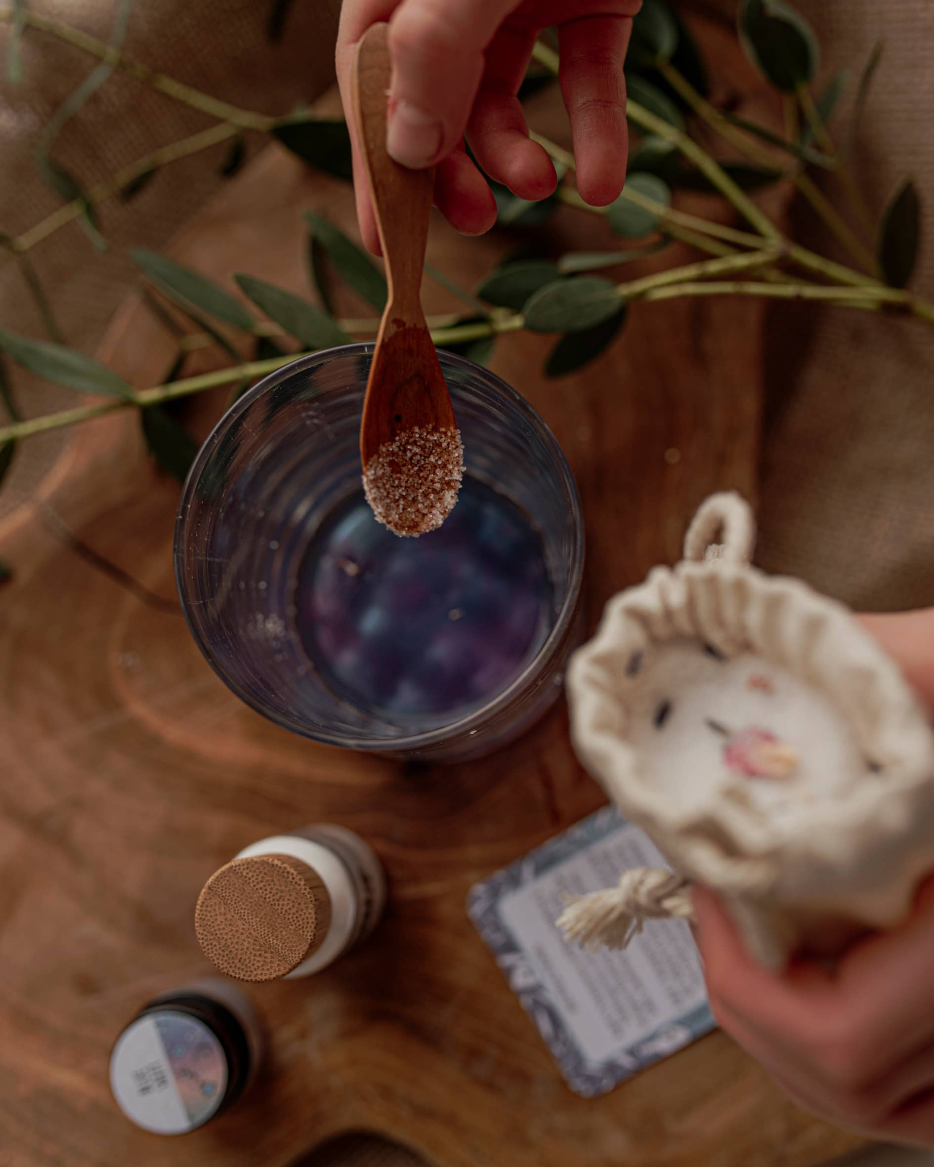 A person's hand is holding a wooden spoon with salt over a glass of water, creating a scene reminiscent of THE LITTLE POTION CO's MINI Colour Mood Potion Kit (Colour Changing Kit). Another hand grips a small cloth bag. Additional items, including a bottle and a small container, rest on the wooden surface with green leaves in the background.