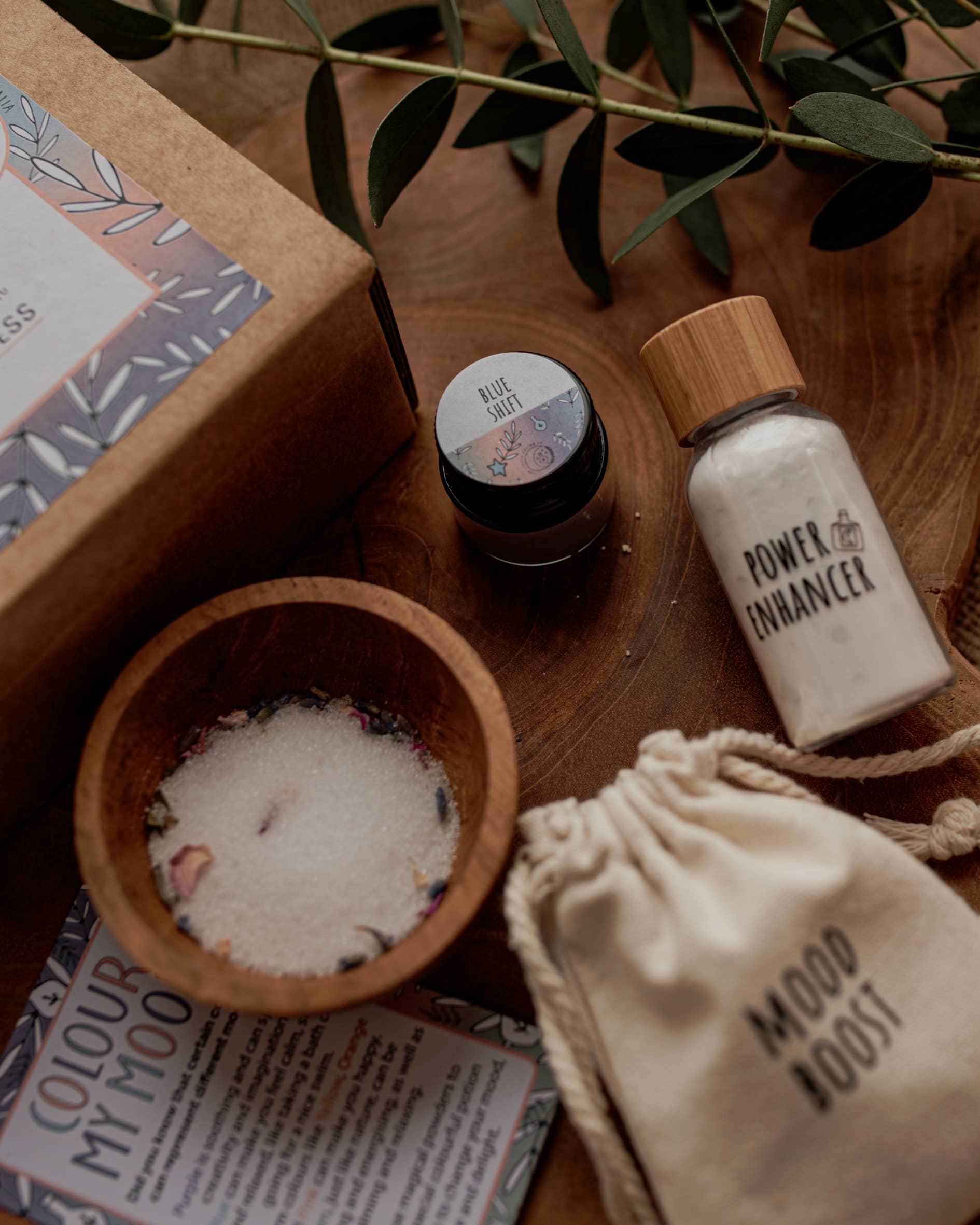 A flatlay of various wellness items on a wooden surface, featuring a small jar labeled "Blue Bath," a glass bottle labeled "Power Enhancer," a drawstring bag labeled "Mood Boost," a bowl of bath salts, and a card labeled "Color My Mood," all reminiscent of the delightful **MINI Colour Mood Potion Kit** by **THE LITTLE POTION CO** for sensory play.