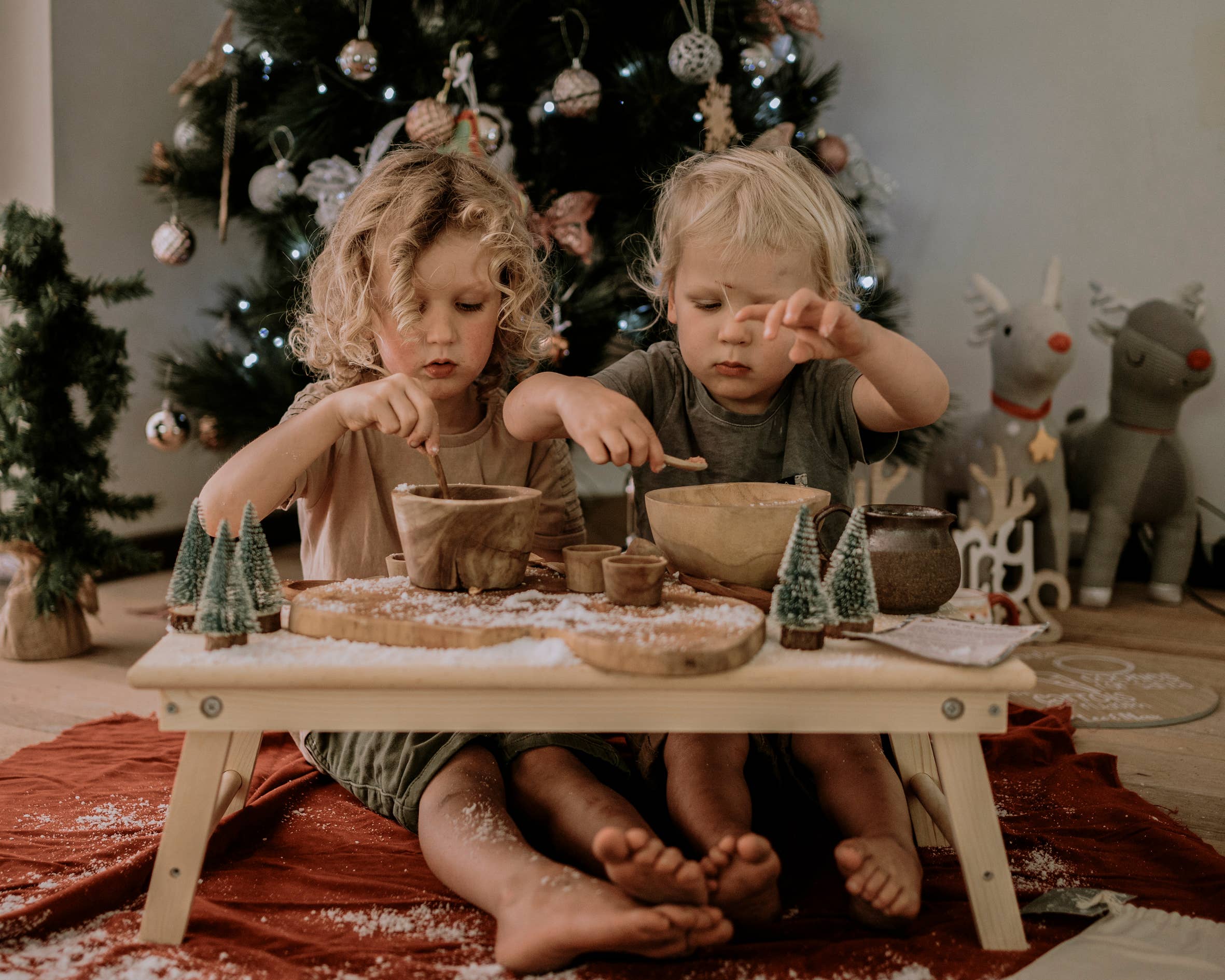 Two young children sit on a red blanket, enjoying the Xmas Eve Potion Pouch to Call the Reindeer from THE LITTLE POTION CO on a small wooden table. In the background, a Christmas tree adorned with lights and ornaments glimmers beside decorative reindeer, creating a scene that is cozy and filled with magical tradition.