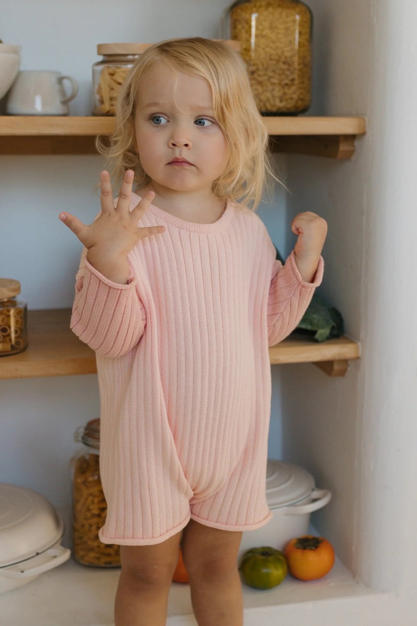 A young child with blonde hair stands in a kitchen wearing a Golden Knit Romper Strawberry Sundae from GOLDEN CHILDREN. They are looking to the side with one hand raised near their face and the other resting on the counter. Shelves with jars of pasta and a pot are in the background.