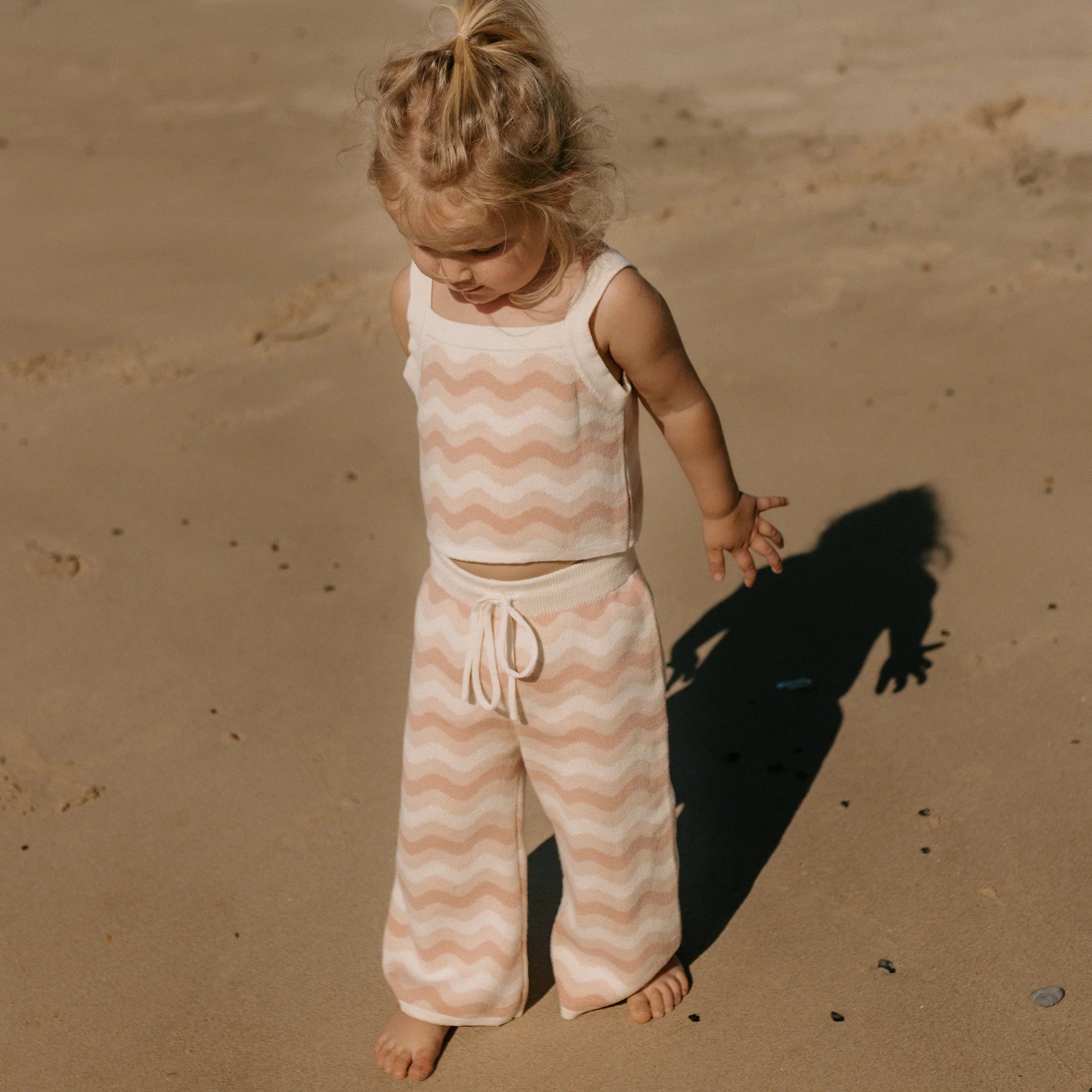 A toddler with blonde hair stands on a sandy beach wearing the Seashore Knit Set Strawberry Cream by GOLDEN CHILDREN, featuring a sleeveless top and matching wide-leg pants adorned with a pink and white wavy pattern. The seaside-inspired ensemble casts gentle shadows as the child gazes down at the sand.