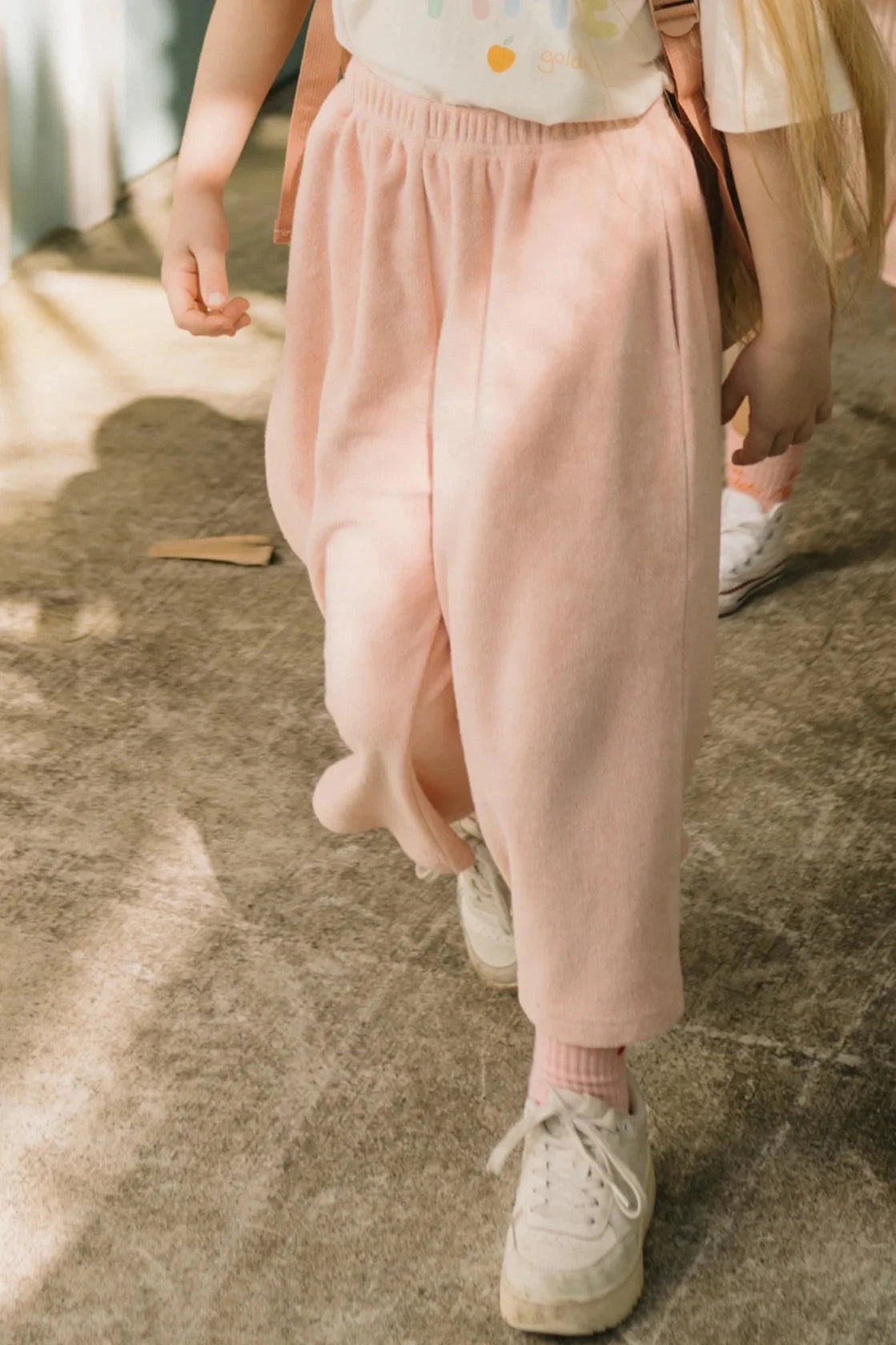 A child wearing GOLDEN CHILDREN's Confetti Wide Leg Pants in Pink Parfait and white sneakers strolls on a sun-dappled concrete path, capturing the charming pink parfait tone of their attire. Another child in light clothing is partially visible behind, adding to the scene. Shadows from nearby objects create a confetti-like pattern on the ground.