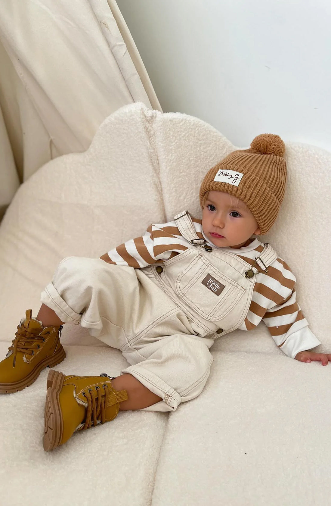 A young toddler wearing the Camden caramel striped pullover and creme overalls and boots with the pom pom beanie in rusty.