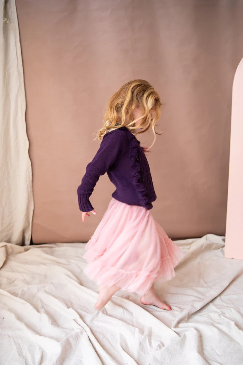 A young girl twirling in the carrie tutu in the pink lady apple colour. She is wearing a purple knitted sweater and standing on cream drapes.