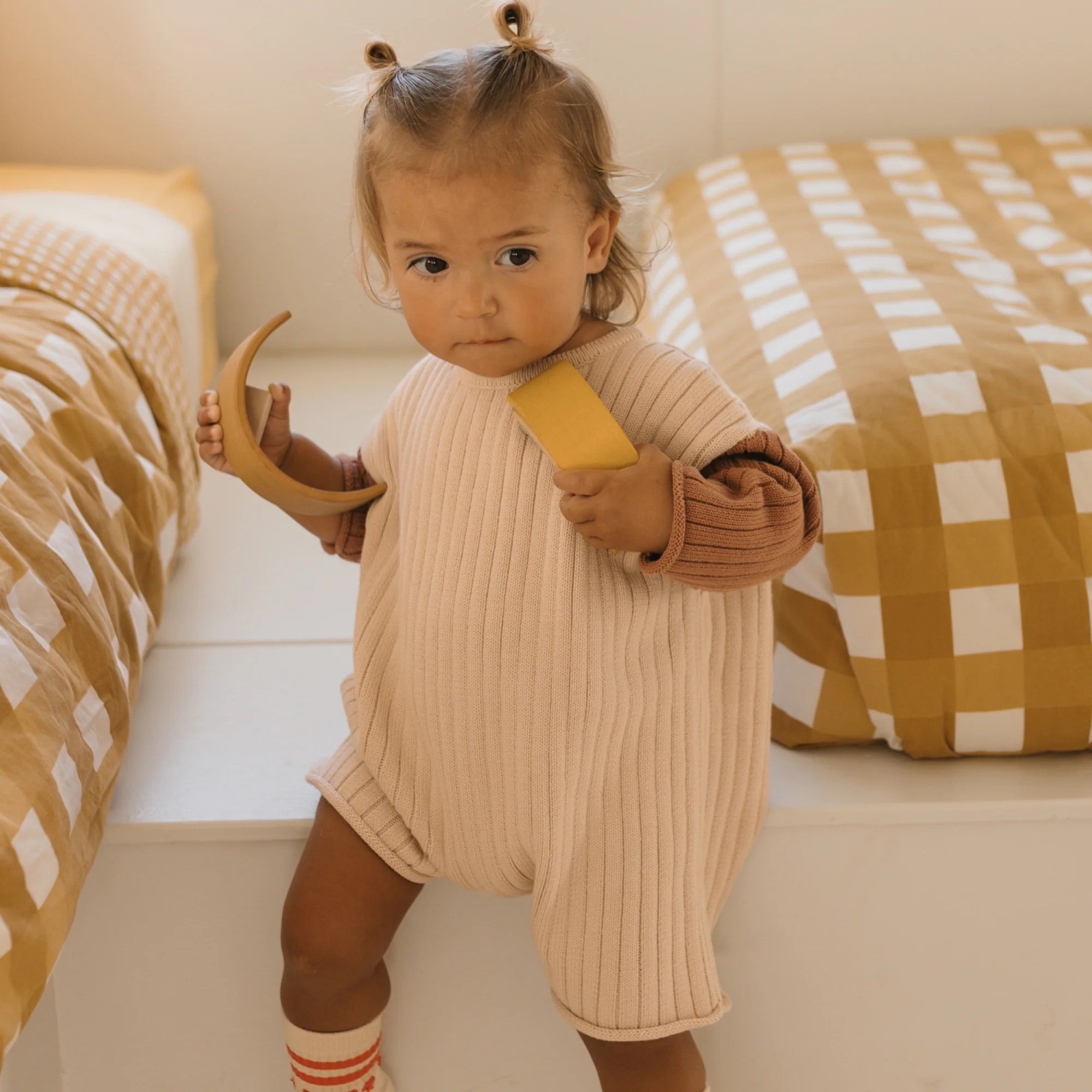 A toddler with light brown hair styled in two small buns is standing between two beds with a plaid pattern in yellow and white. The child is wearing the Golden Knit Romper Caramel Sundae by GOLDEN CHILDREN over a pink long-sleeved shirt and holding a wooden toy moon and block.