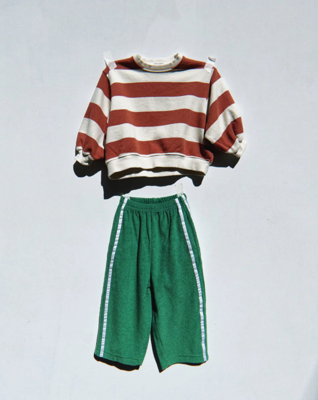 The child's outfit, set against a neutral background, includes a 100% terry cotton, long-sleeved sweater with brown and white stripes and buttoned shoulder straps, paired with ZIGGY ZAZA's Ziggy Zaza ~ Twin Stripe Terry Pant Verde in green, featuring an elastic waist for comfort.