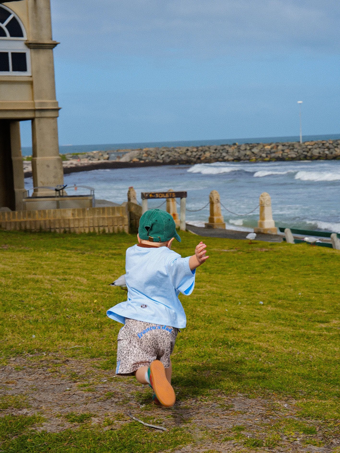 A child in a blue shirt and oversized PRE-ORDER Leopard Cargo Shorts by FRANCO'S DAD runs towards the ocean on a grassy area. The child is wearing a green cap and facing away from the camera. The sea and a stone structure are visible in the background under blue skies.