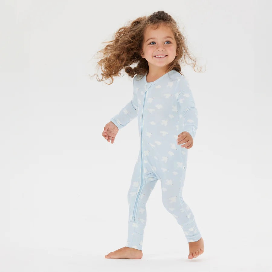 A smiling child with curly hair walks barefoot in the Day or Night Onesie Bird Song from KYND BABY, a light blue baby onesie crafted from EcoCosy® Viscose. This sustainable fabric showcases white designs, set against a plain white background.