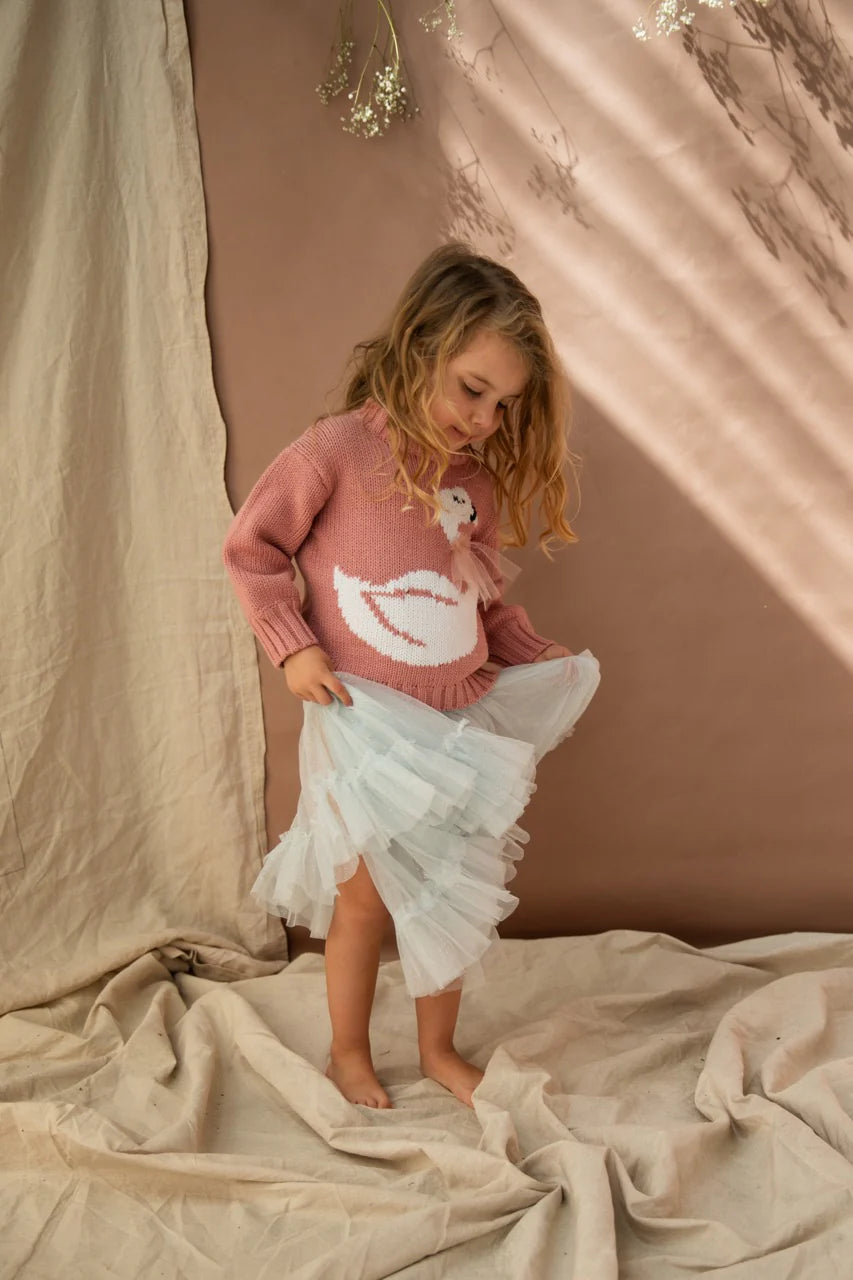 A young girl wearing the carrie tutu in iced lake colour with a knitted jumper with a duck on it. She is in a room with brown drapes.