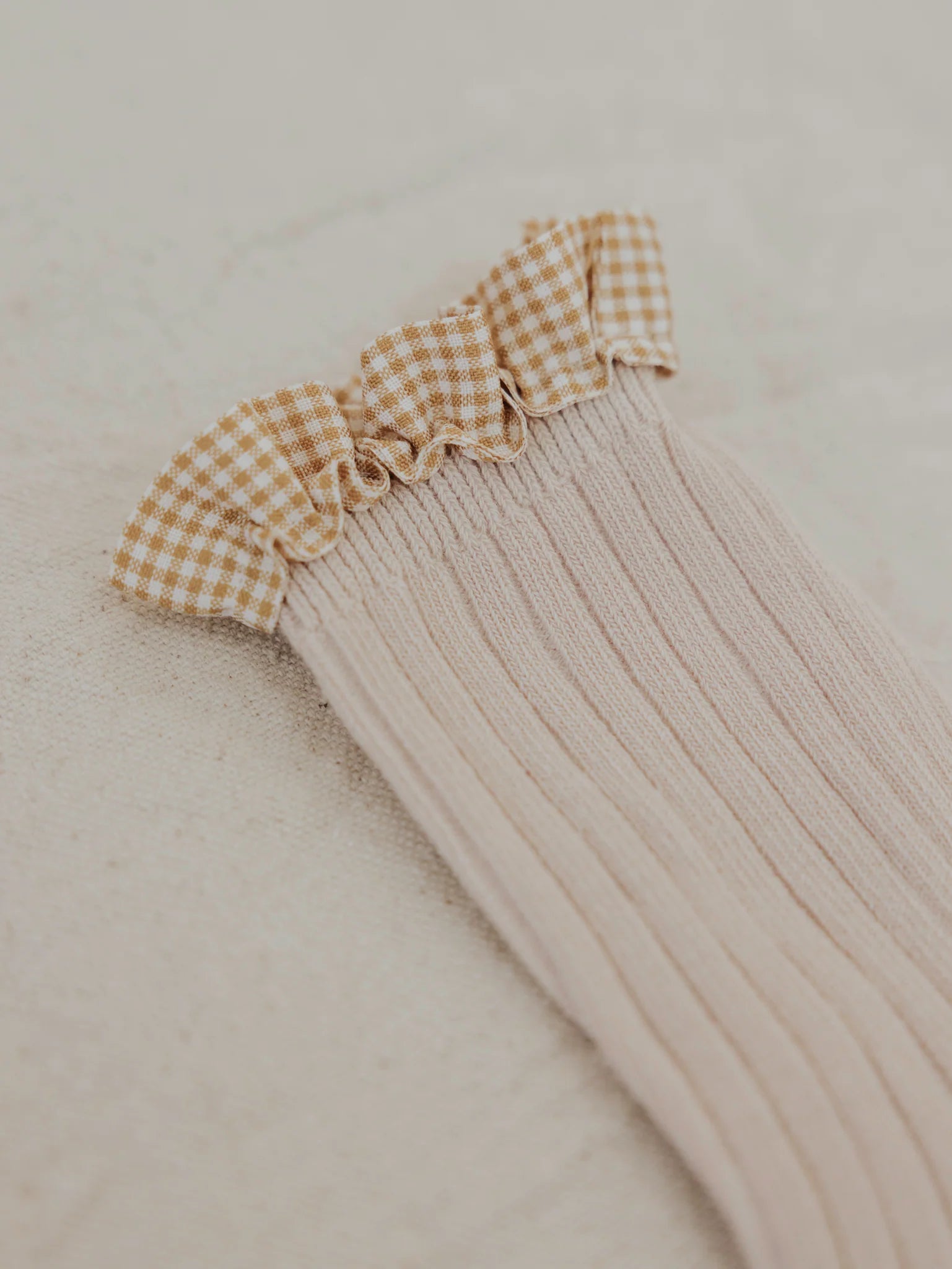 The Frilly Socks Oat by VALENCIA BYRON BAY are made of ribbed beige fabric with a ruffled edge adorned in a white and mustard checkered pattern, all laid out on a light background.