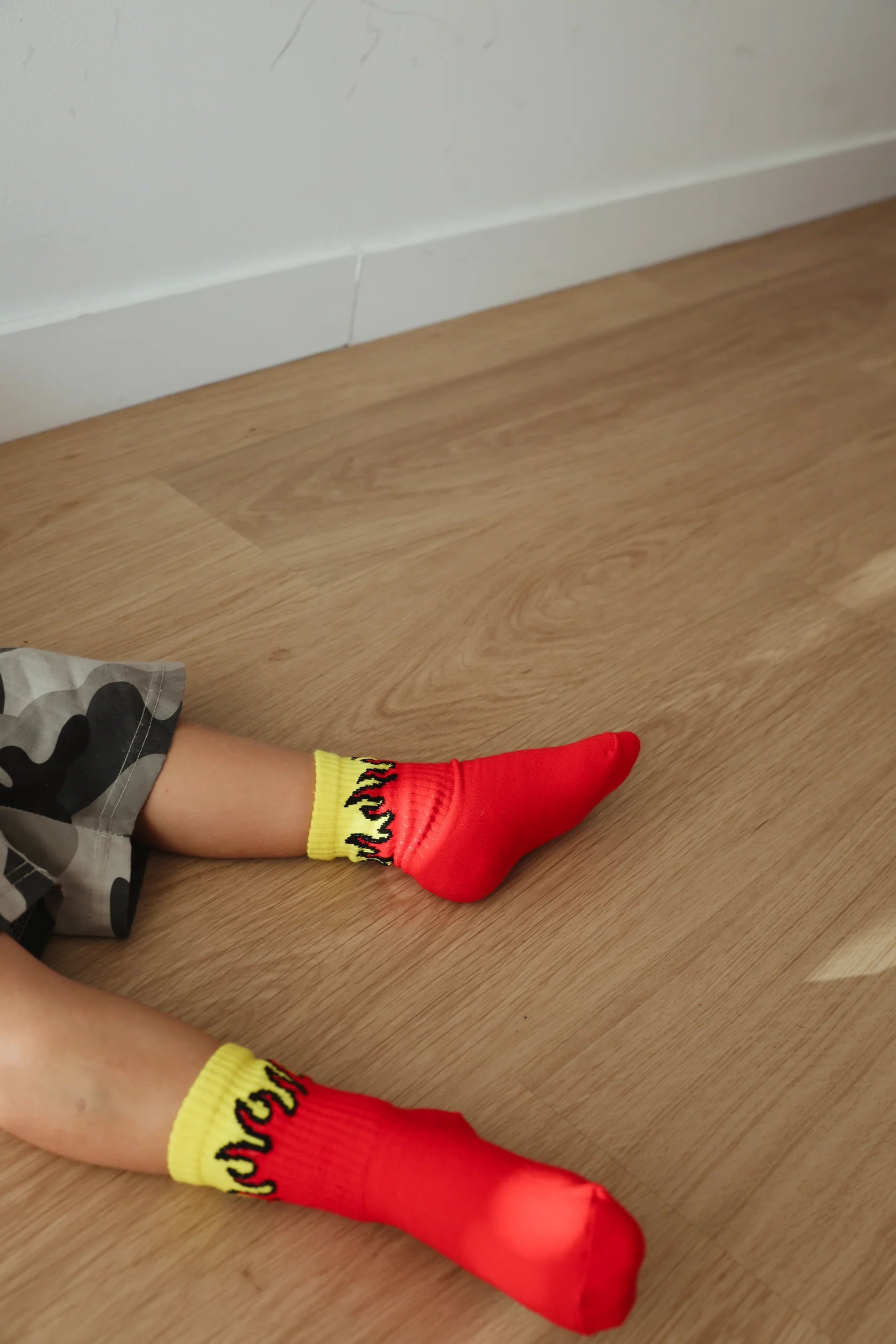 A child, dressed in gray camouflage shorts with elastic banded ankles peeking out, sits on a light wooden floor wearing TINY LOVE CLUB's Fire Sock—a pair of tiny red socks featuring a yellow trim and black flame designs. The corner of a white wall is visible in the background.