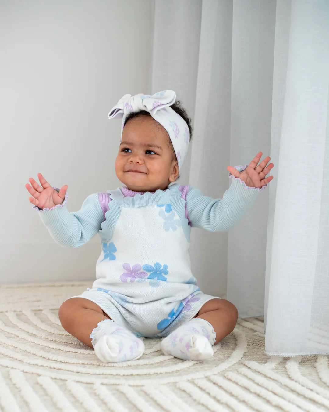 A baby sits on a patterned carpet wearing the Playsuit Alaska by ZIGGY LOU, made of 100% cotton Jacquard knit and adorned with pastel purple and blue flowers. The baby also wears a matching headband and socks, with hands raised and a cheerful expression. White curtains are seen in the background, adding to the serene setting.