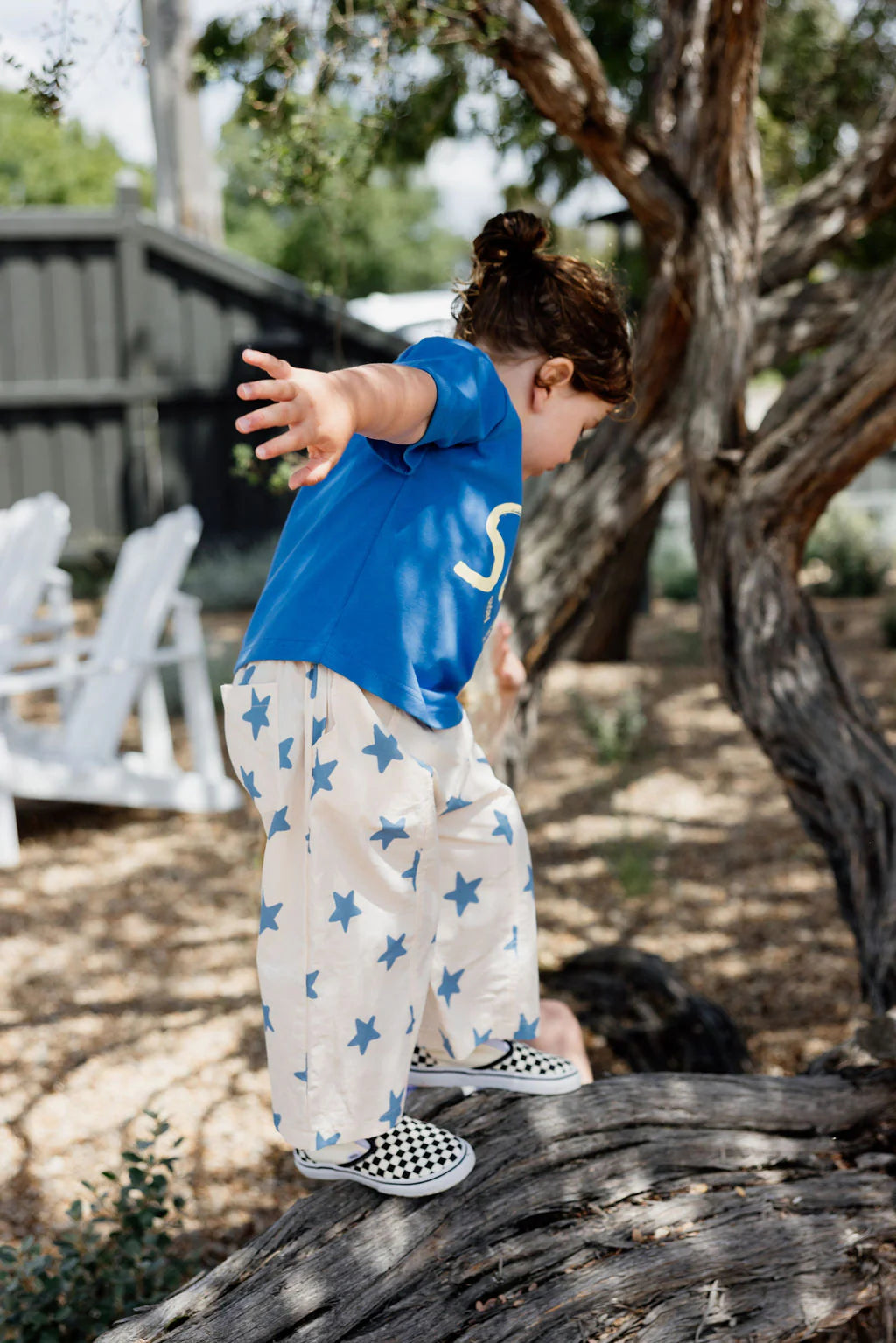 A child in a blue shirt and SUNDAY SIBLINGS' Dusty Star Pants, featuring an elastic waistband, balances on a fallen tree trunk. They're wearing checkered shoes and stretching their arms for balance against a backdrop of trees and blurred outdoor seating.