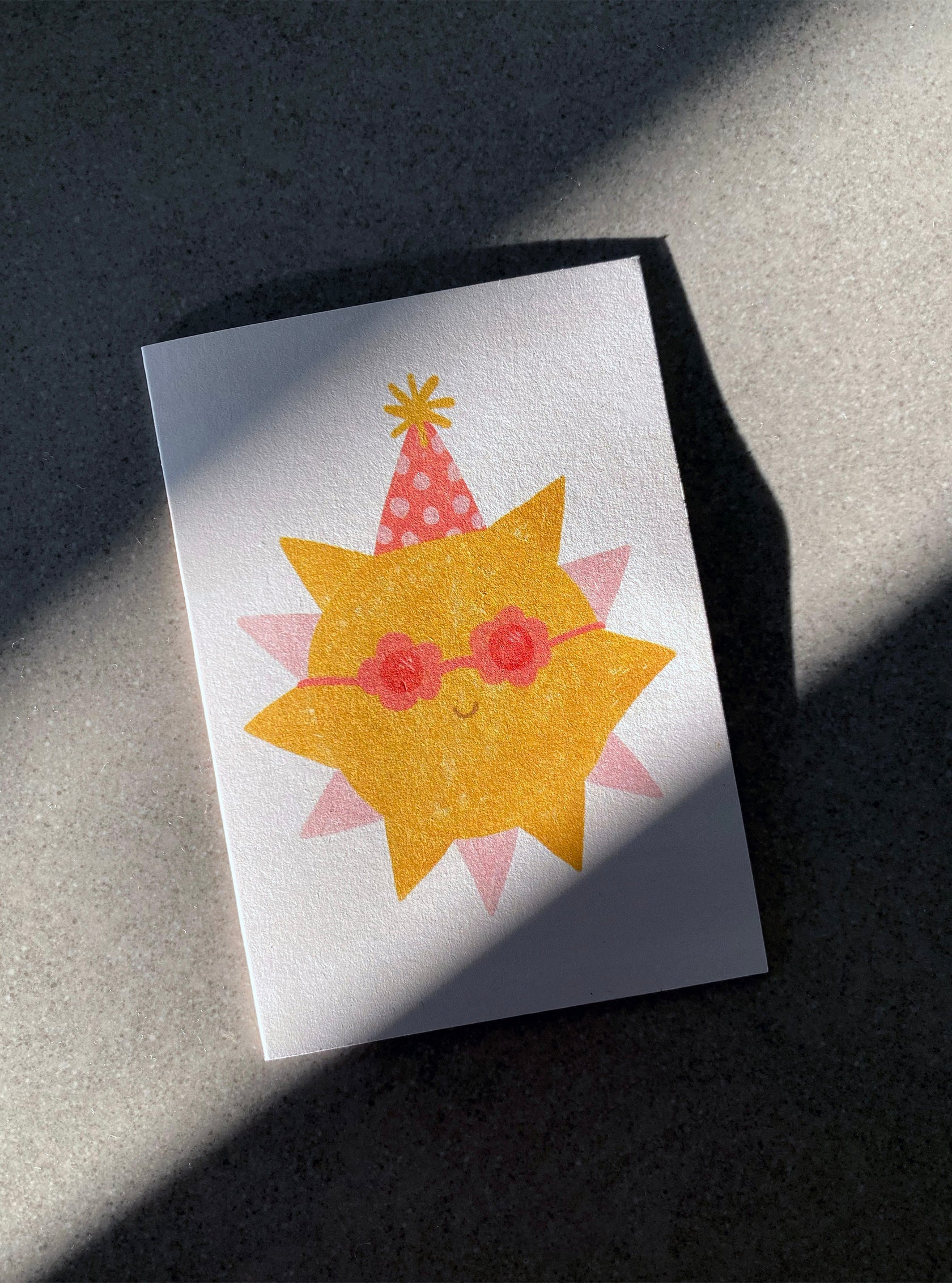 The Sunshine Mini Card by LAUREN SISSONS STUDIO features a cheerful yellow sun wearing red sunglasses and a red polka-dotted party hat with a yellow pom-pom. The sunrays are illustrated as a pink halo around the sun on a grey surface, complete with shadows cast across the recycled paper—making it an ideal addition to any birthday celebration.