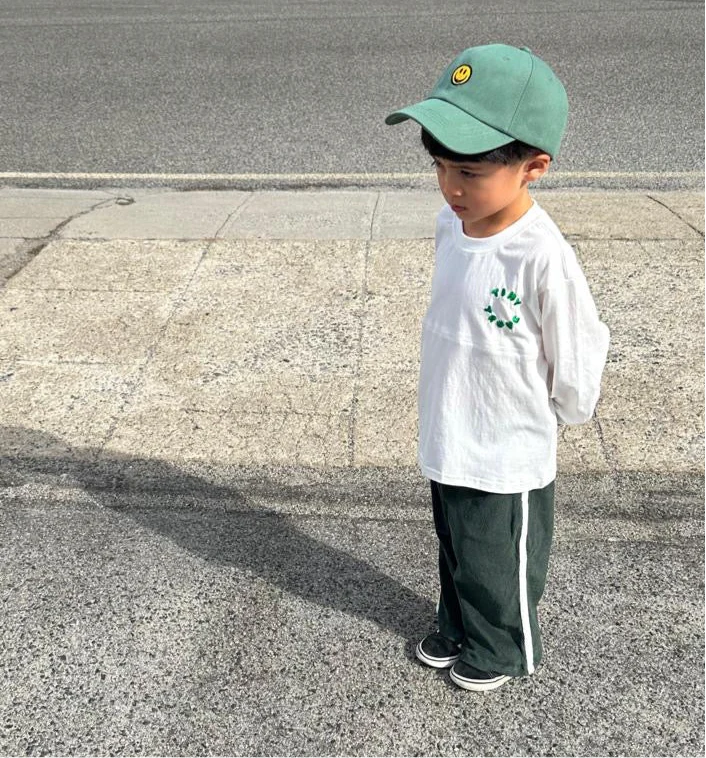 A small child stands on the sidewalk looking down, wearing a green baseball cap with a yellow smiley face, a white long-sleeve shirt with a leafy design, Darcy Racer Pants Moss by TINY TROVE featuring an elastic waistband, and black shoes. The child's hands are clasped behind their back.