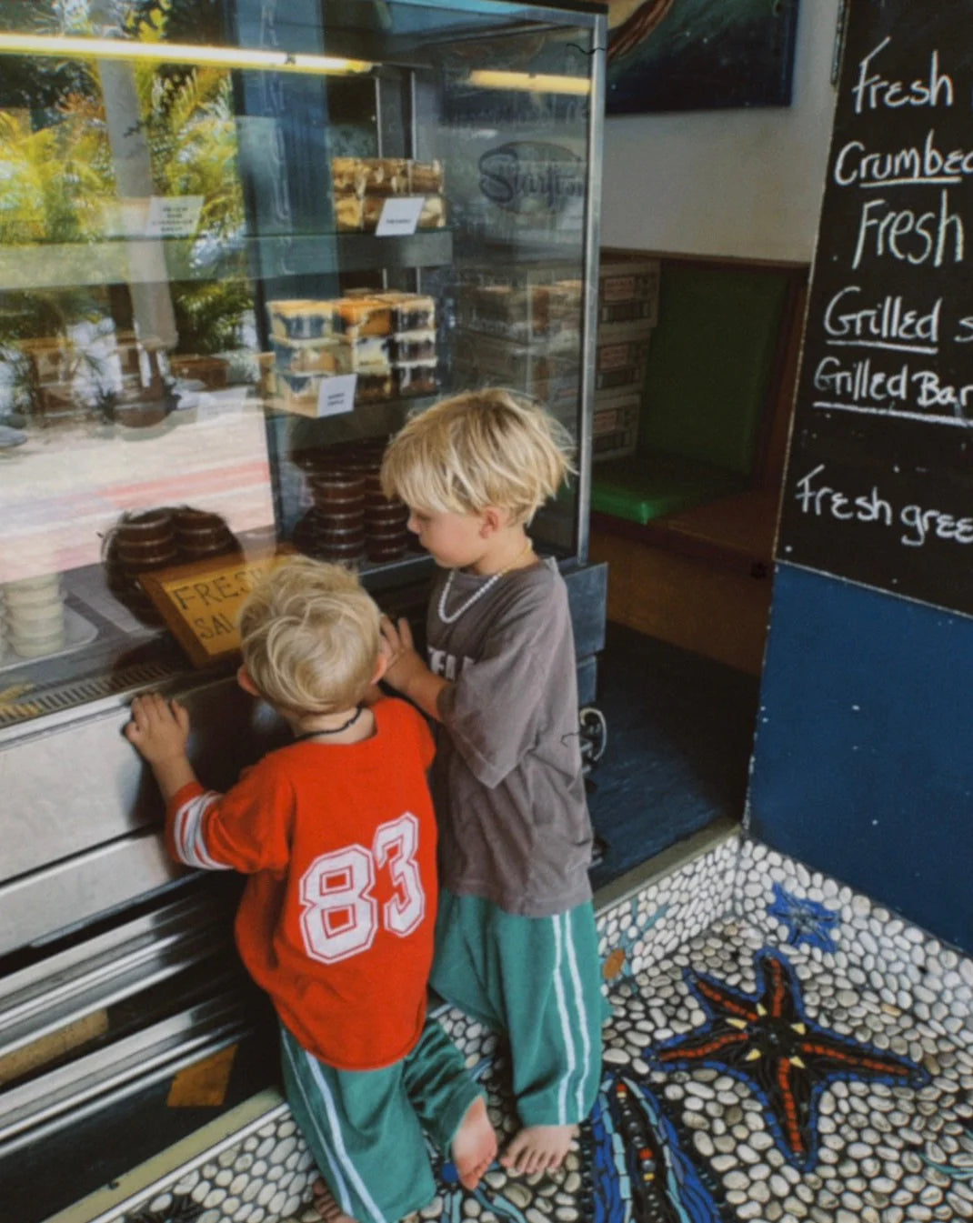 Two young blond-haired children eagerly gaze at baked goods through a glass display. One child in a red shirt with the number 83 wears Ziggy Zaza ~ Twin Stripe Terry Pant Verde, and the other is in a gray shirt. They stand on a starfish mosaic floor near a chalkboard menu.