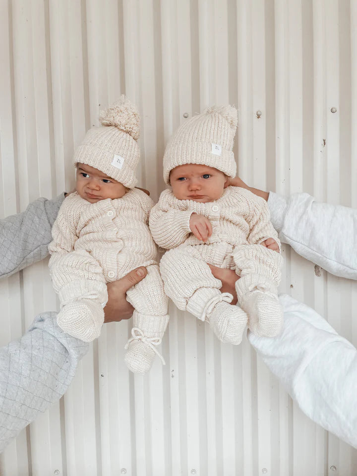 Two infants are held up by two pairs of hands against a white, ridged background. Both babies are wearing matching Classic Knit Romper Honey outfits from ZIGGY LOU, complete with pom-pom hats in a creamy beige color. The textured cotton knit of their rompers adds a cozy charm. They are looking at the camera, and their expressions vary slightly from curious to calm.