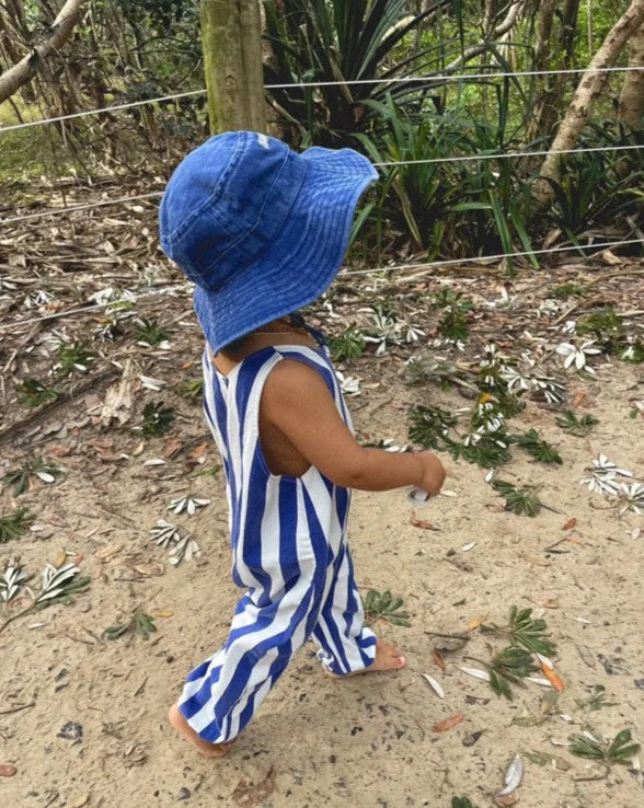 A toddler strolls along a sandy path wearing the Ziggy Zaza Everyday Scout Hat Blu and blue-and-white striped overalls. The scenery includes greenery and plants, with leaves scattered on the ground.