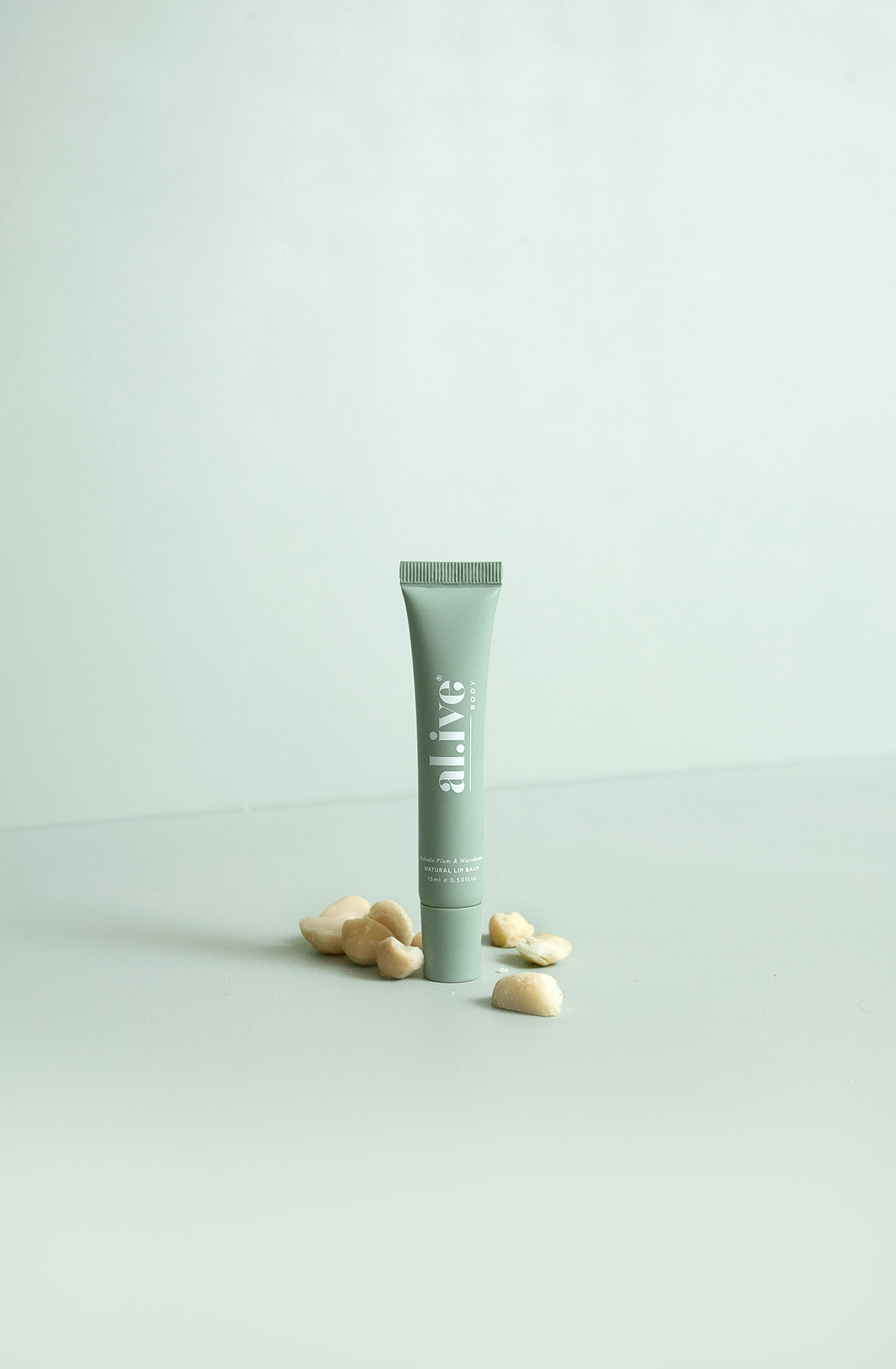 A green squeeze tube of al.ive natural lip balm with kakadu plum and macadamia surrounded by macadamia nuts on a green surface and background.