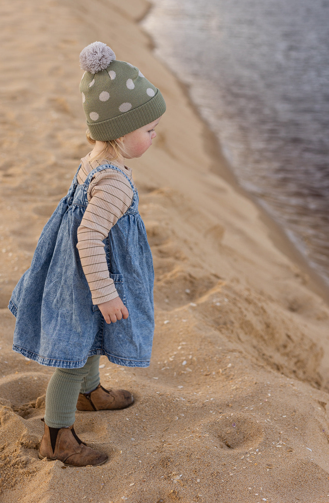 A young girl on the beach looking out to the water wearing the raindrops beanie in sage by acorn kids.