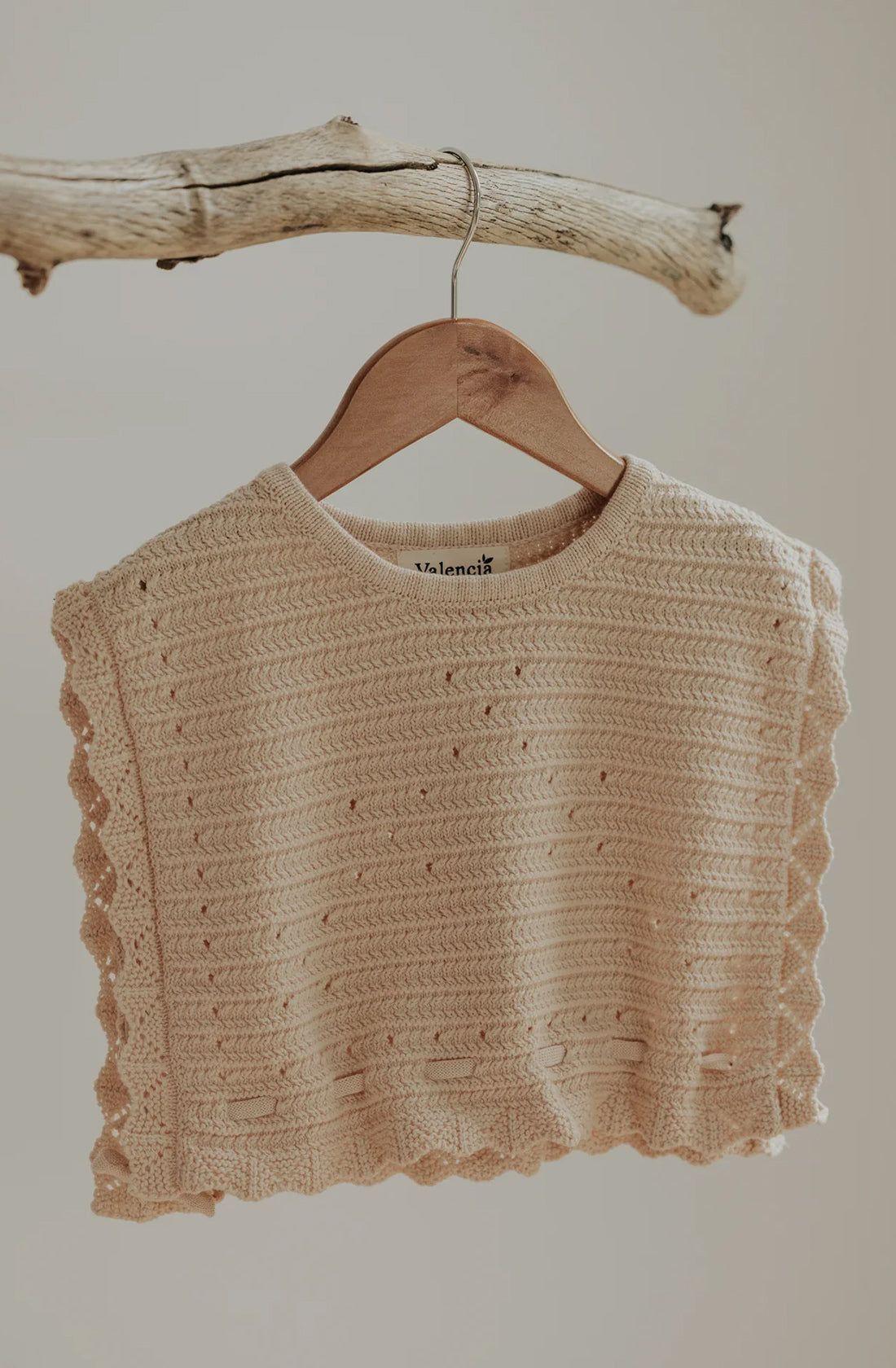 A beige Grove Vest by VALENCIA BYRON BAY, made of crocheted cotton with a lace-like, scalloped edge, is hanging on a wooden hanger hooked onto a rustic piece of driftwood. This cropped top, in a warm neutral tone, features small, evenly spaced holes and an intricate design—perfect as an effortless layering piece.