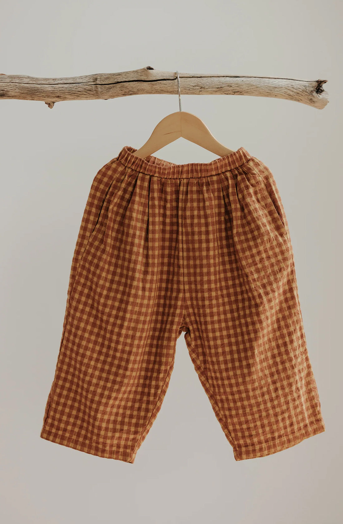 A pair of Rio Pant Honeycomb by VALENCIA BYRON BAY, crafted from 100% sand-washed cotton in a unisex design, hangs from a wooden hanger suspended on a rustic tree branch against a plain beige background. These loose-fitting, brown and beige checkered pants feature an elastic waistband and exude a relaxed, comfortable appearance.