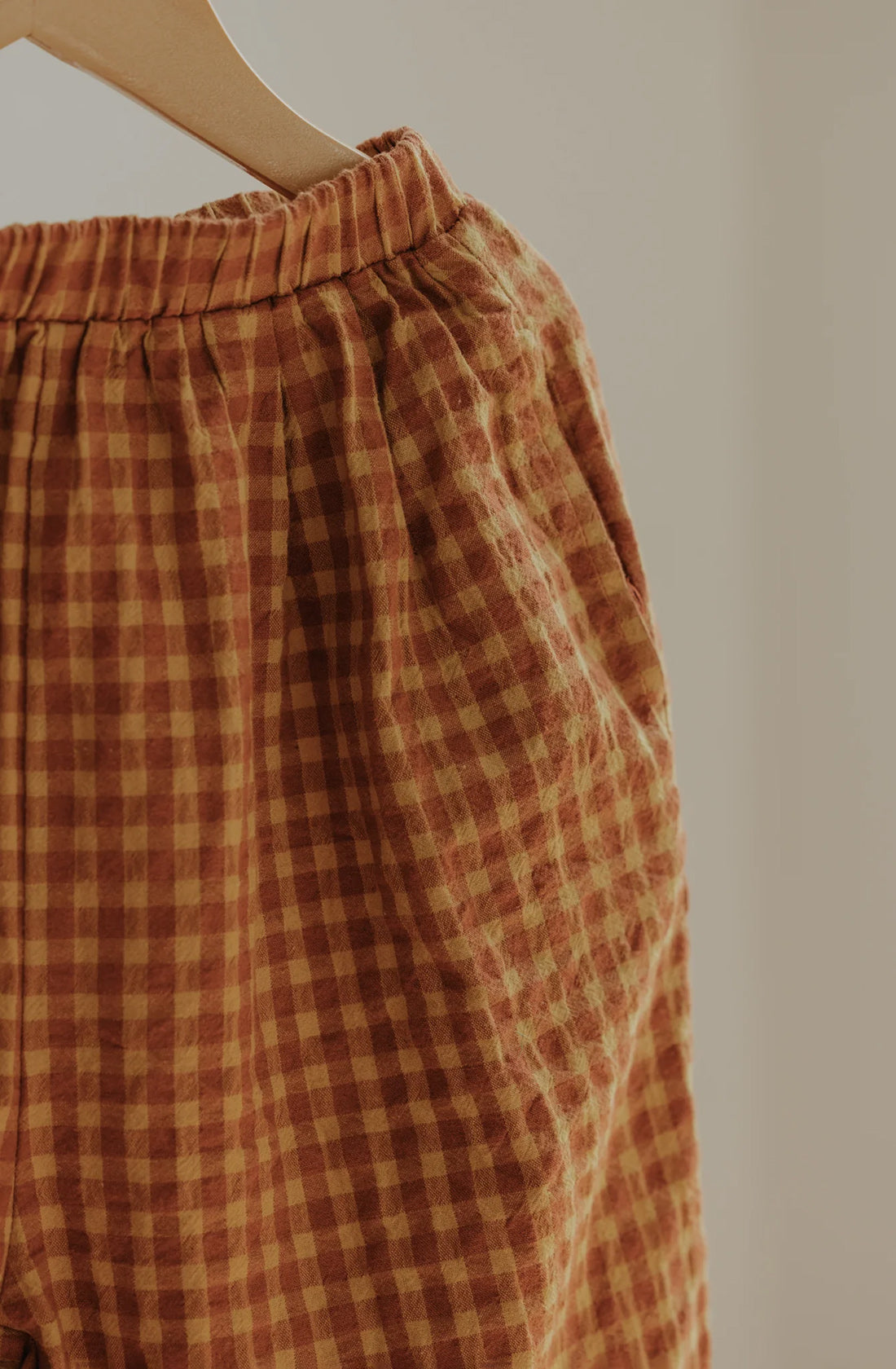 A close-up of the Rio Pant Honeycomb by VALENCIA BYRON BAY in rust and beige gingham checkered pattern on a hanger, showcasing the elastic waistband and 100% sand washed cotton fabric. The unisex design is emphasized against a neutral background, highlighting the texture and details of the pants.